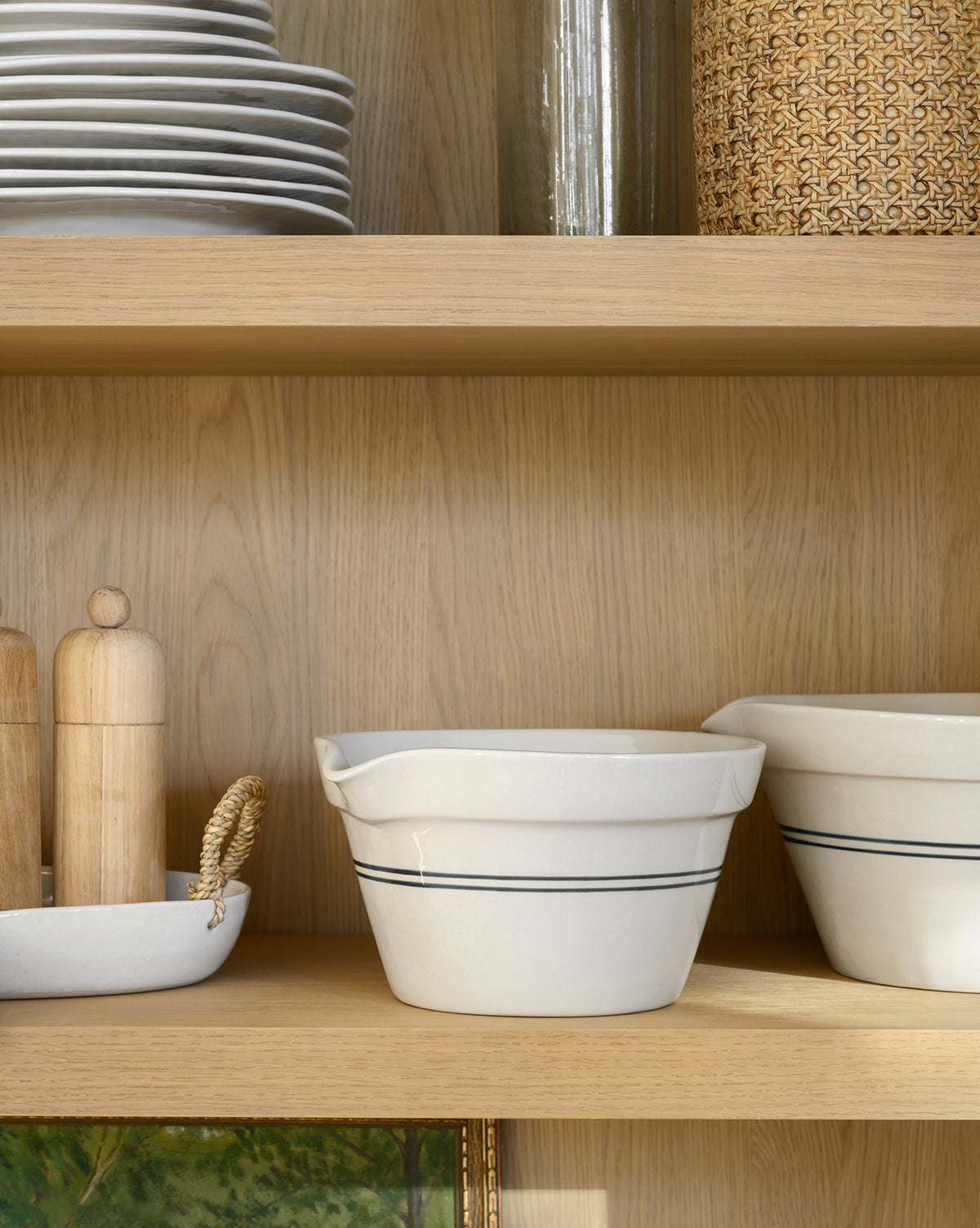Everett Mixing Bowls with black stripes rest on a wooden shelf next to a dish with wooden salt and pepper grinders; plates and woven and glass containers are displayed on the shelf above, by McGee & Co.