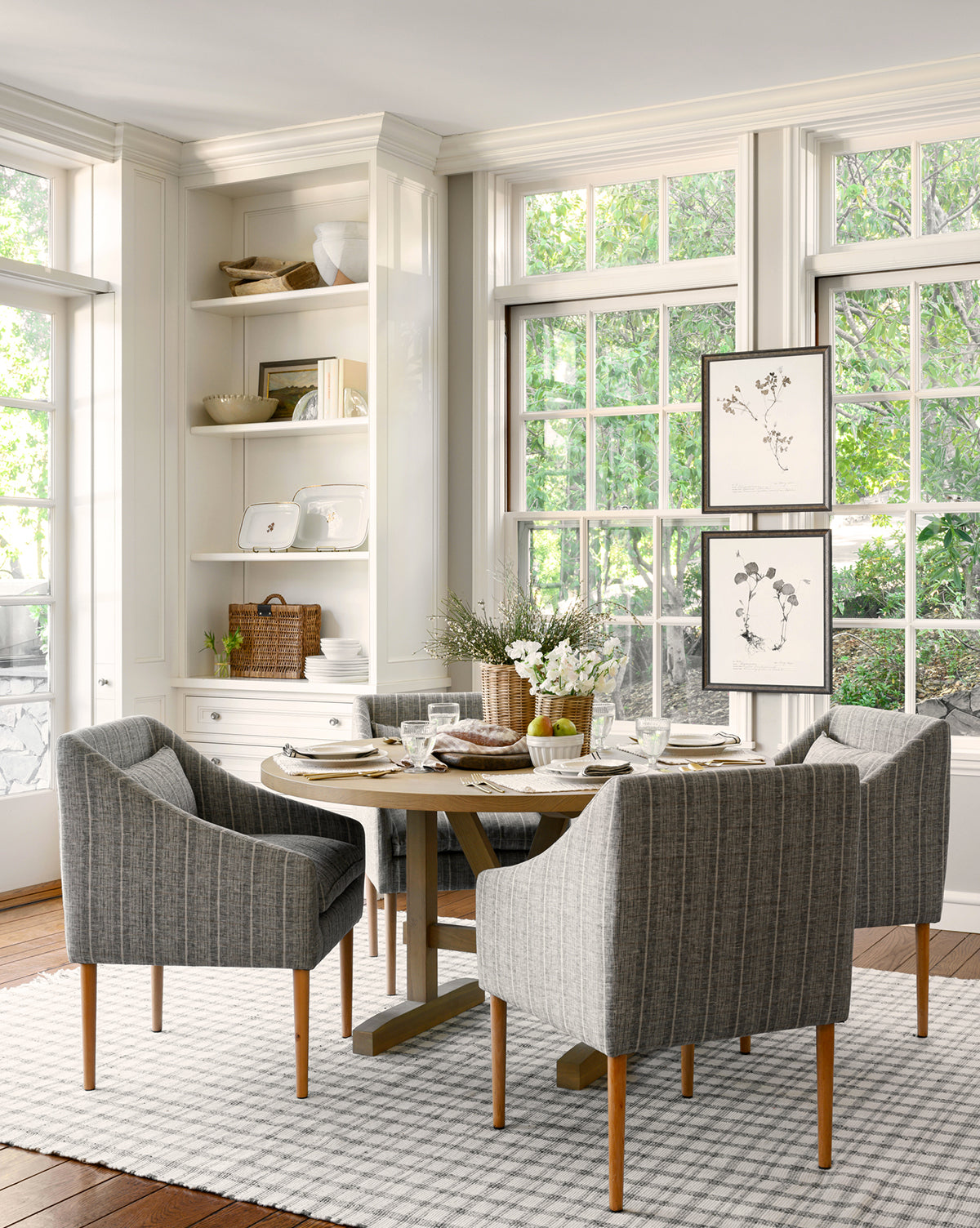 A bright dining area features a round wooden table, four gray upholstered chairs, a striped rug, built-in shelves with decor, large windows with tree views, and the Subdued Botanicals II wall art adding charm to the space.