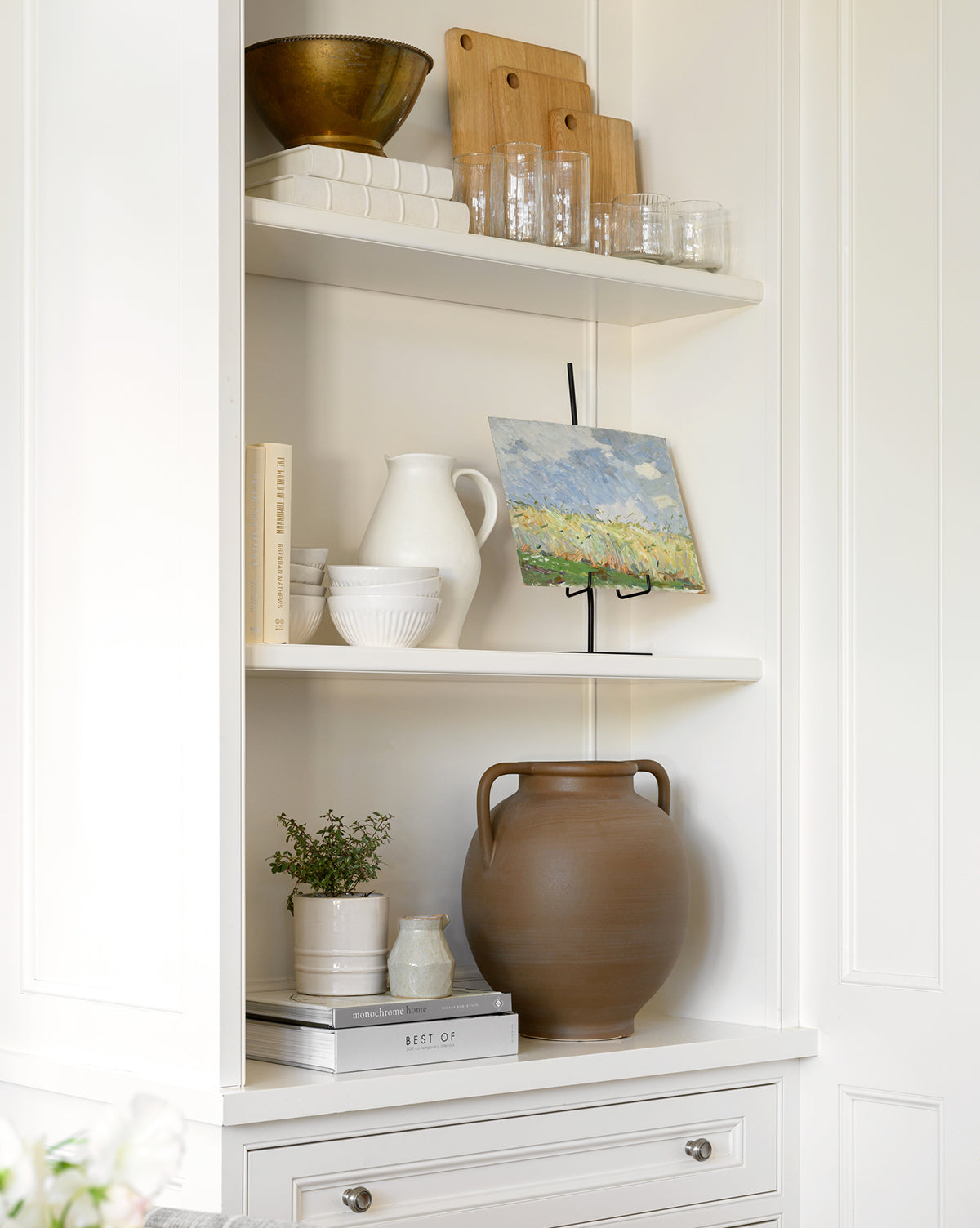 A white built-in shelf displays glassware, cutting boards, a bowl, books, white ceramic pitchers, small plants, a large brown vase, and a small landscape painting on a Metal Easel.