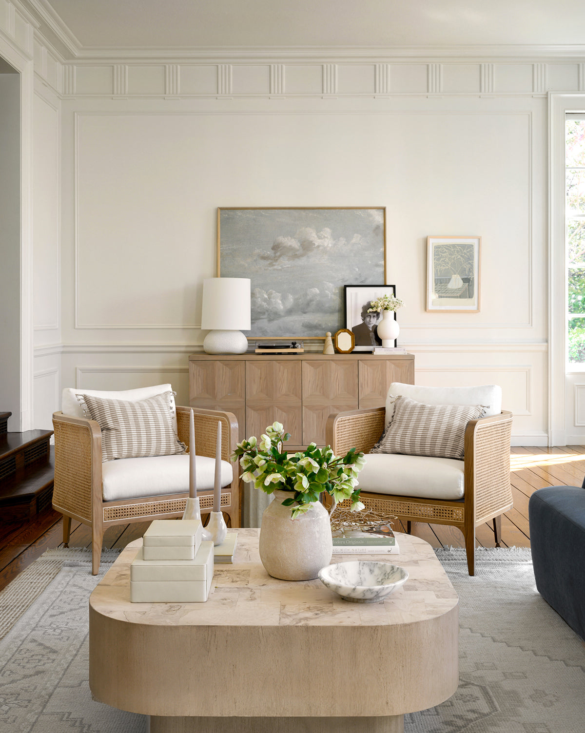 A bright, modern living room with two Elowyn Lounge Chairs featuring natural caning and white cushions, a light wood coffee table with flowers, a sideboard topped with art and lamps, and a large cloud painting above the sideboard, by McGee & Co.