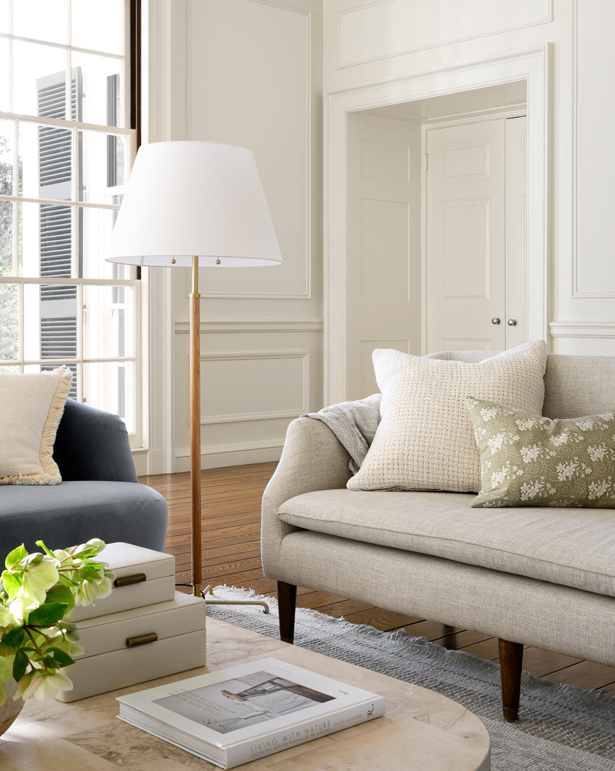 A bright living room features a beige sofa, floral and knitted cushions, a blue armchair, the stylish Florence Floor Lamp, a coffee table with books and plants, wood floors, and large windows that fill the space with natural light, by McGee & Co.