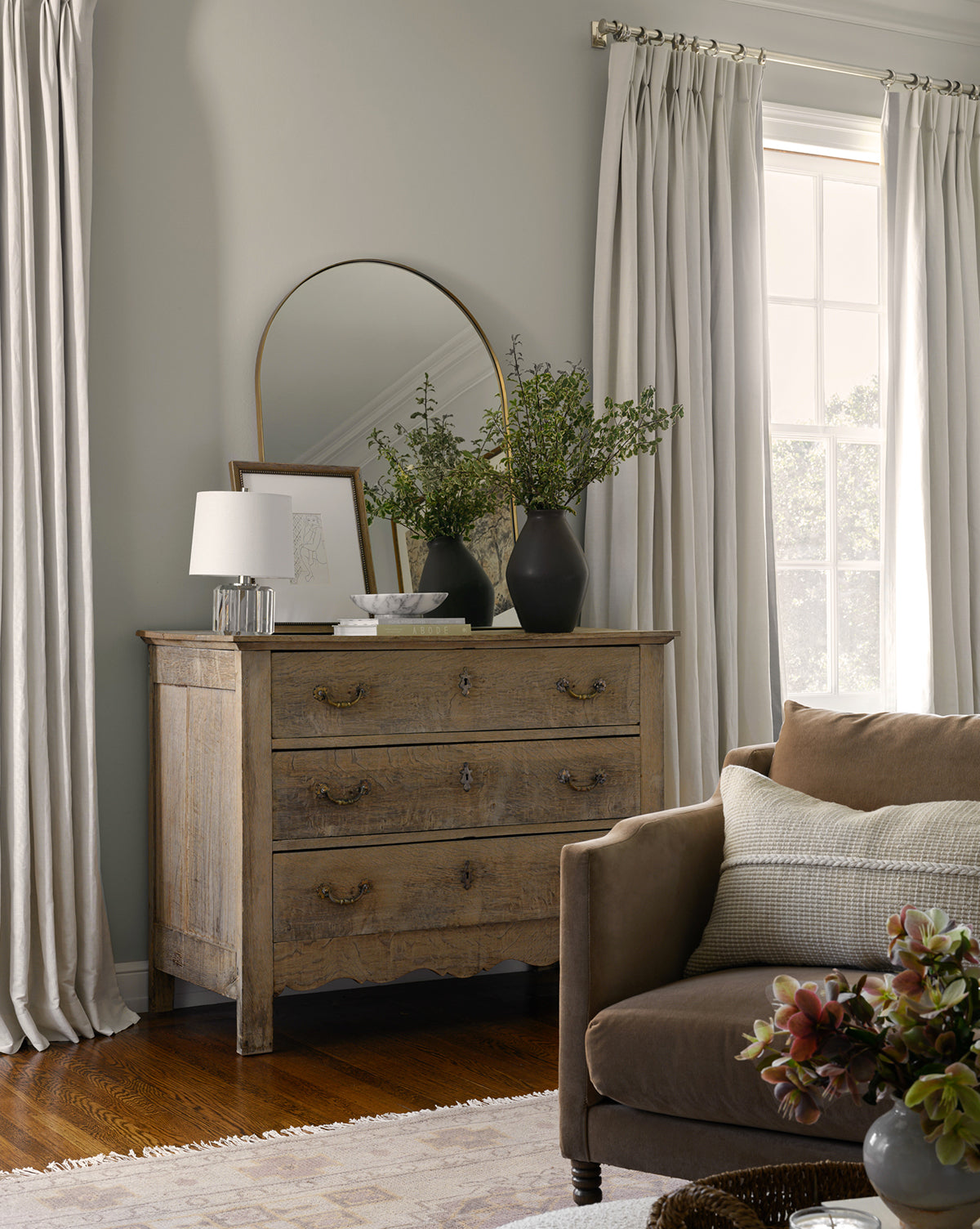 A cozy living room corner with a brown sofa, a wooden dresser topped with greenery vases, the Wylie Arched Mirror featuring shatterproof glass, framed art, and a white lamp. Light curtains frame a sunlit window for an inviting feel, by McGee & Co.