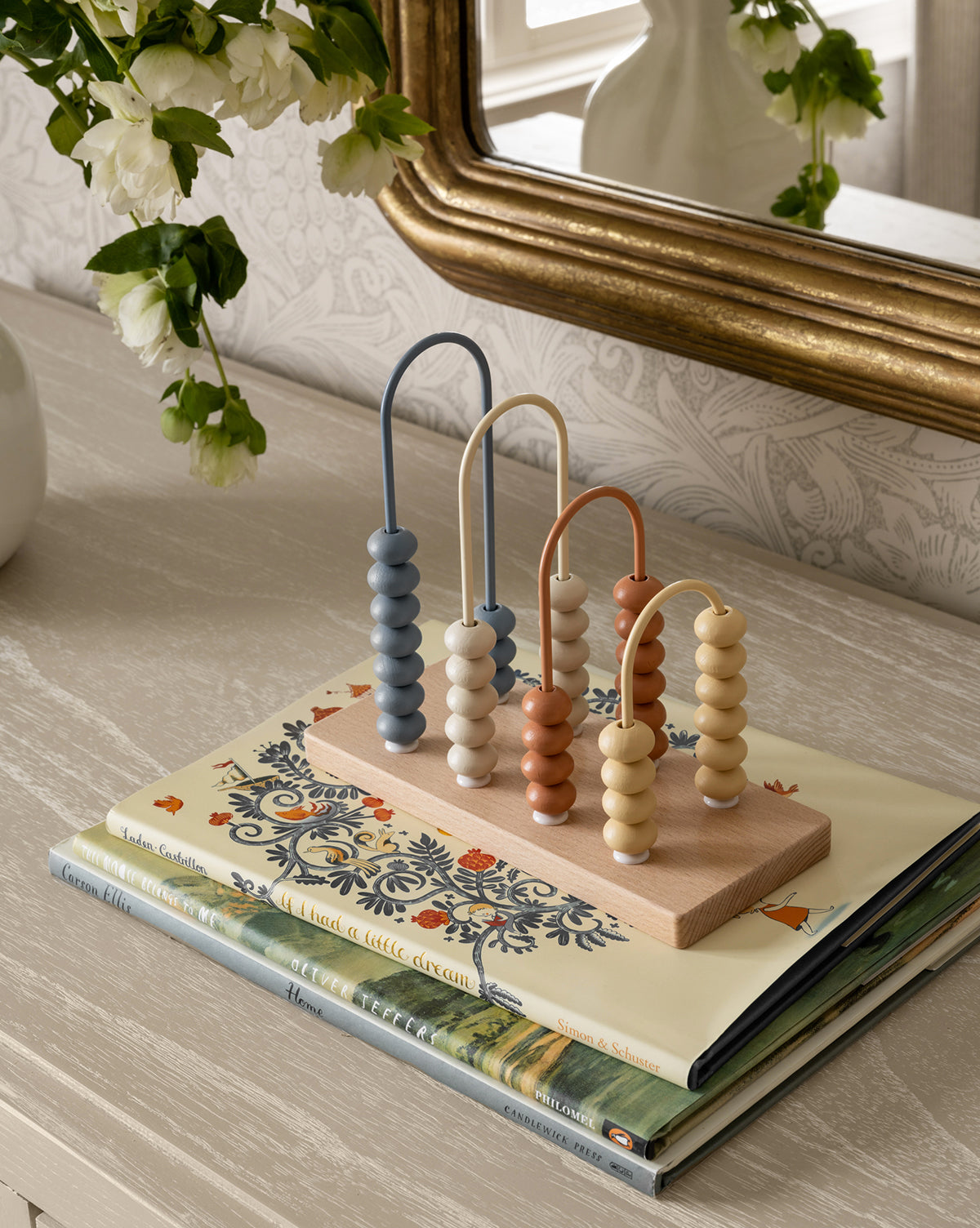 A small wooden bead maze toy with pastel beads sits on top of three stacked books, including This Moose Belongs to Me, on a light wood table beside a mirror and a white vase with green and white flowers.