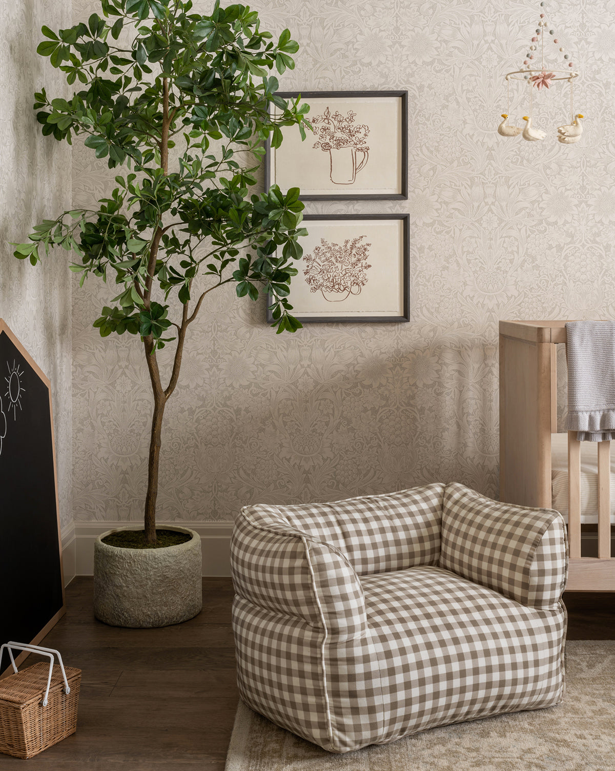 A cozy nursery includes a checkered armchair, a Shady Lady Faux Tree, a wooden crib, neutral decor, framed floral art on the wall, and a small bird mobile hanging from the ceiling.