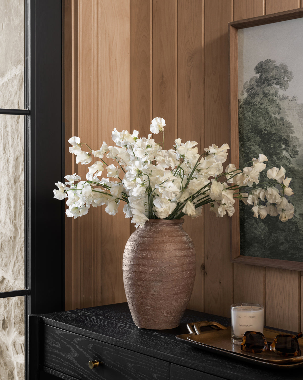 A large brown ceramic vase with white flowers is displayed on a black wooden cabinet beside a candle, sunglasses, and the Adrienne Brass Tray. The backdrop includes wood paneling, a window segment, and a framed landscape painting.