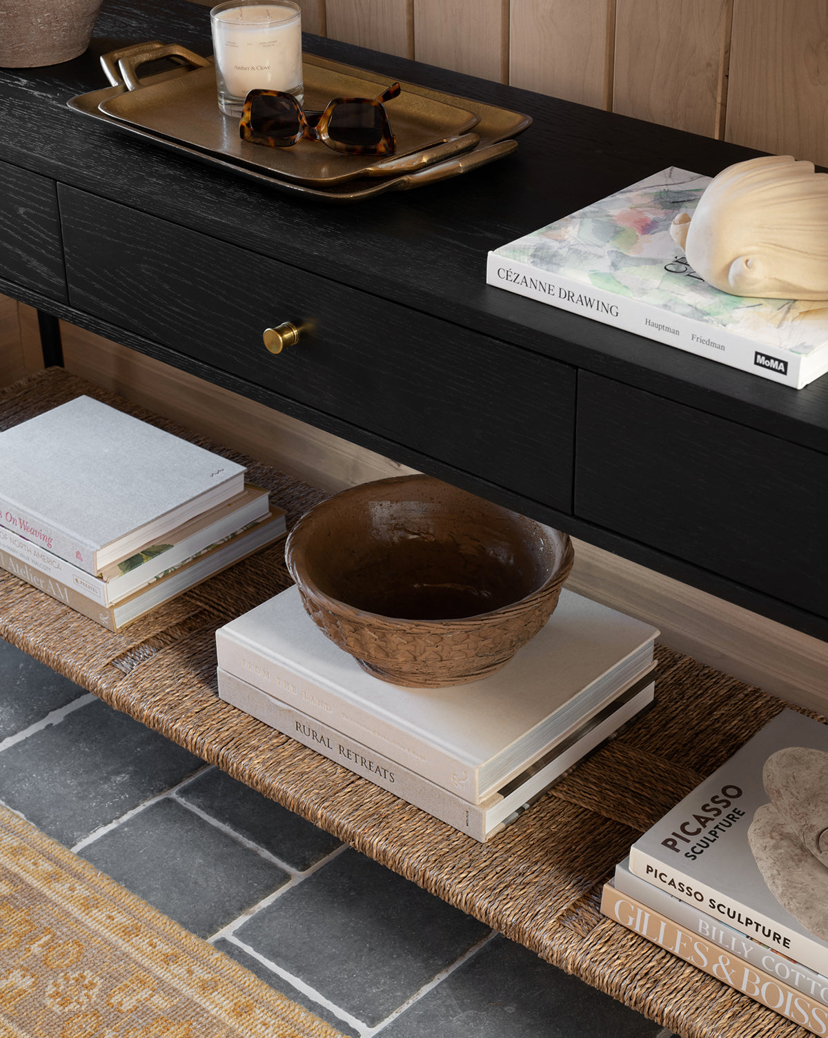 The Dana Console, crafted from modern oak, features stacked art books, a brown ceramic bowl, candles on a brass tray, and a woven lower shelf—ideal for entryway decor—set on a gray stone floor with a golden rug in view, by McGee & Co.