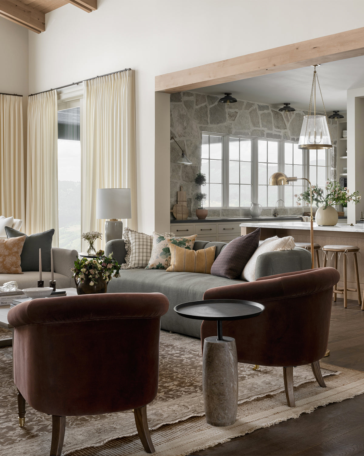 A cozy living room features two burgundy chairs, a gray sofa with pillows, and a stone kitchen backdrop. Neutral tones, soft curtains, and plants surround the Four Hands Sheba Side Table, creating a warm, inviting atmosphere—a scene reminiscent of McGee & Co.