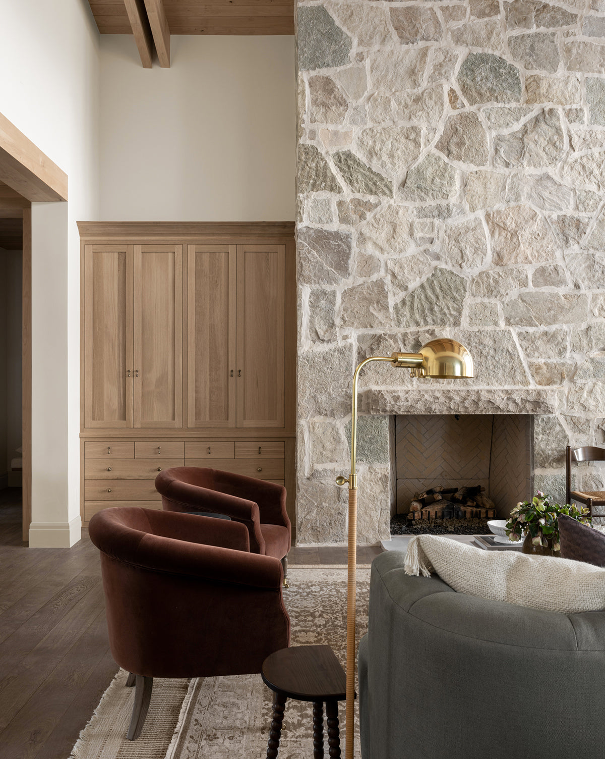 A cozy living room with two brown velvet chairs, a gray sofa, stone fireplace, wooden cabinet, and the Devon Floor Lamp (aged bronze with rattan base), all set on a patterned rug over wooden floors and under exposed beams, by McGee & Co.