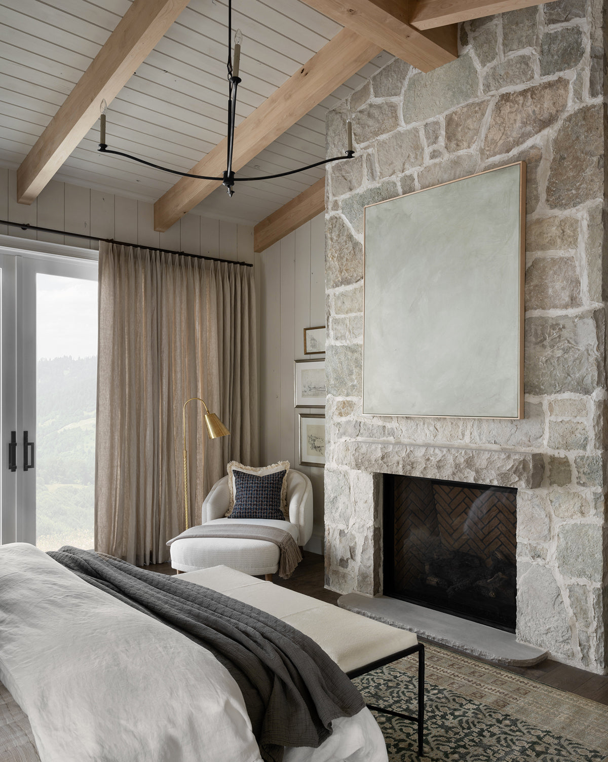 A cozy bedroom with exposed wooden beams, a stone fireplace, and neutral bedding displays the Faded Stone Abstract giclee print above a lounge chair by large windows. Minimalist decor and soft natural light complete this tranquil space.