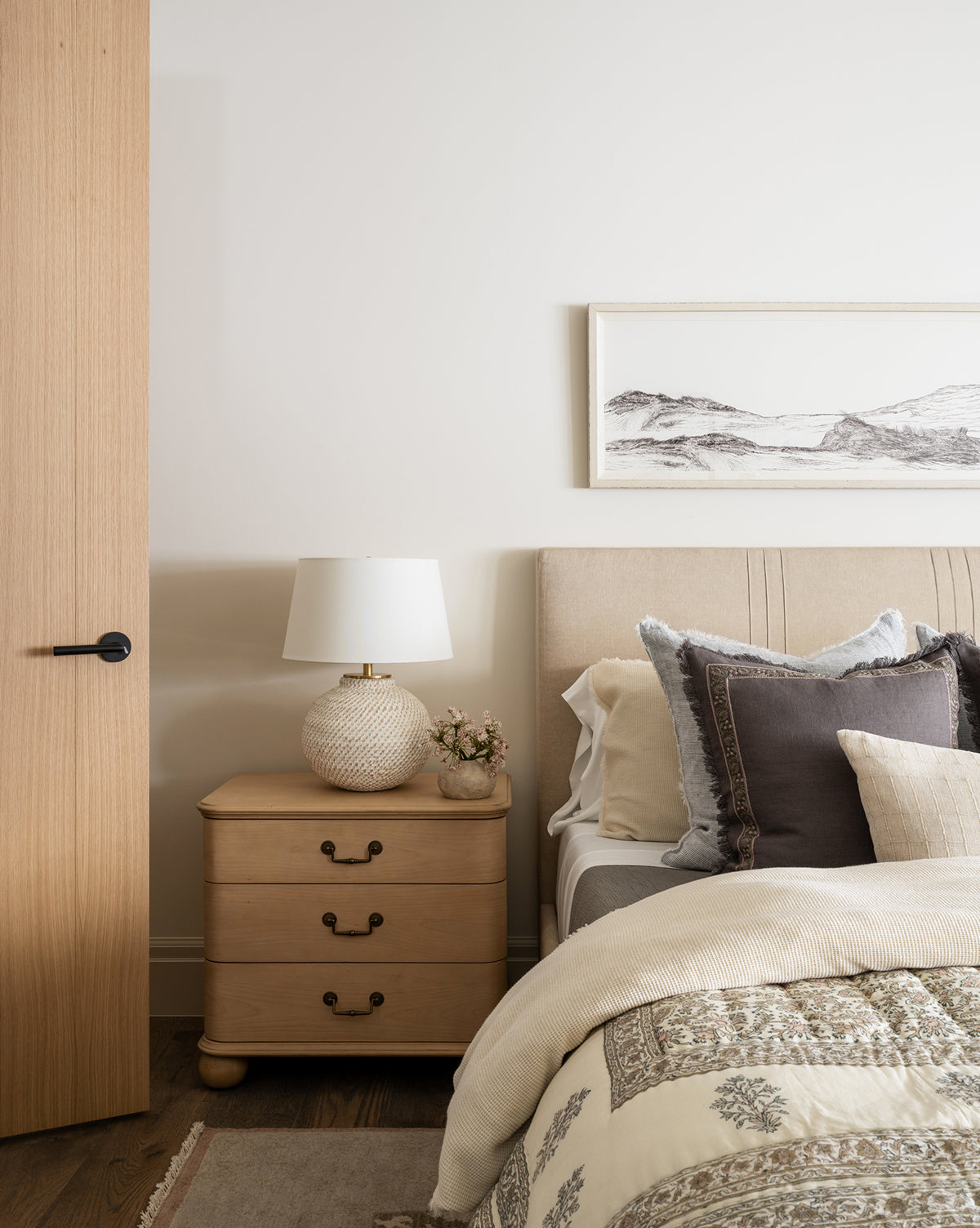 A cozy bedroom corner with a light wood nightstand, the Avedon Table Lamp with a white linen shade, a small vase of flowers, a beige upholstered bed layered with pillows and a patterned comforter, plus minimalist landscape art on the wall by McGee & Co.