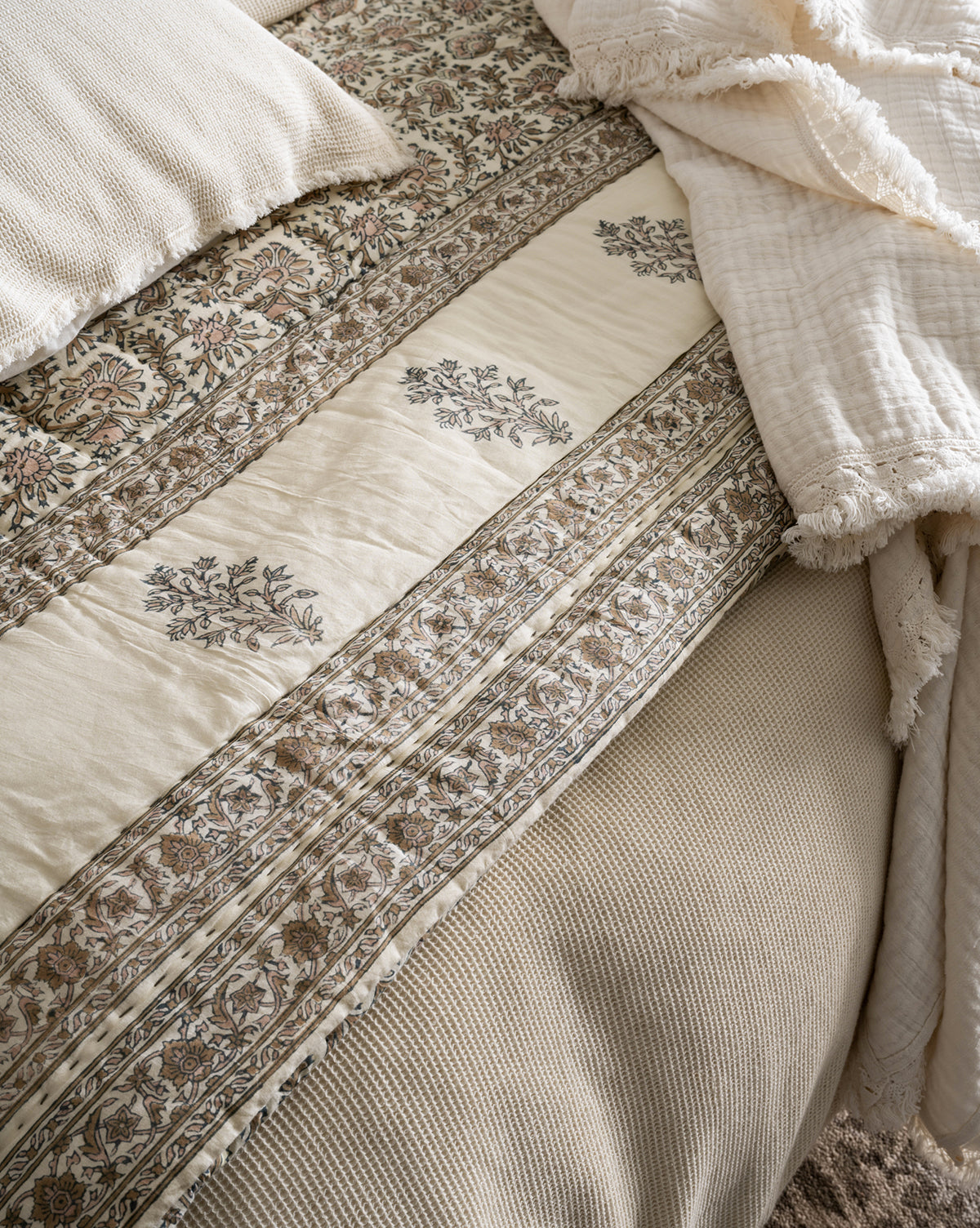A close-up of a bed styled with the Leander Quilt—a cream and beige cotton quilt with intricate floral and botanical patterns—paired with a textured beige blanket and matching pillow for an inviting, cozy look.