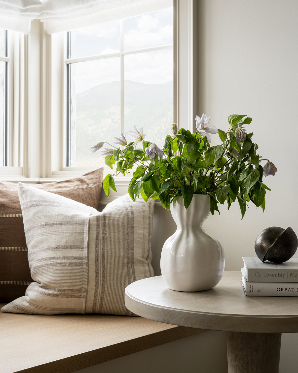 A cozy window seat with striped beige cushions sits beside a round table. On the table, sunlight illuminates stacked books, a white vase with green branches and flowers, and the modern Jackson Offset Sphere decorative object.
