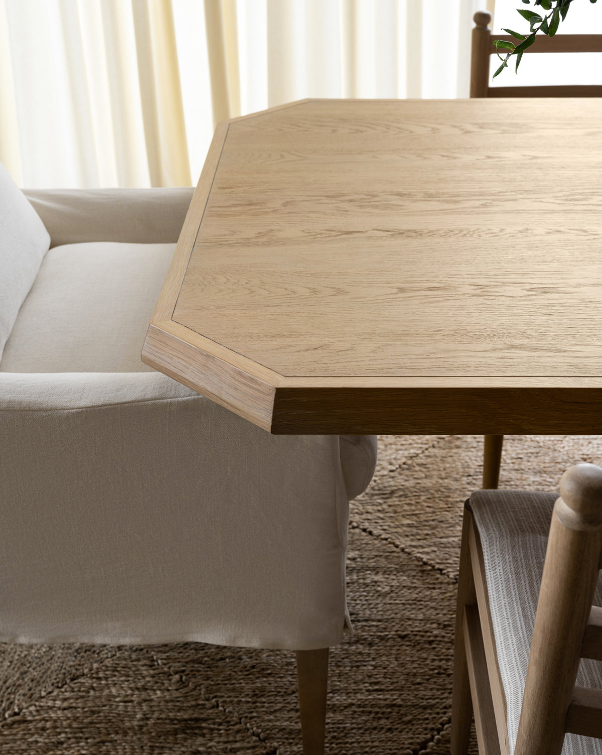 A close-up of the Orville Extension Dining Table with an angled edge, paired with a white upholstered armchair and a wooden chair with a striped cushion, set on a textured rug by sheer curtains, by McGee & Co.