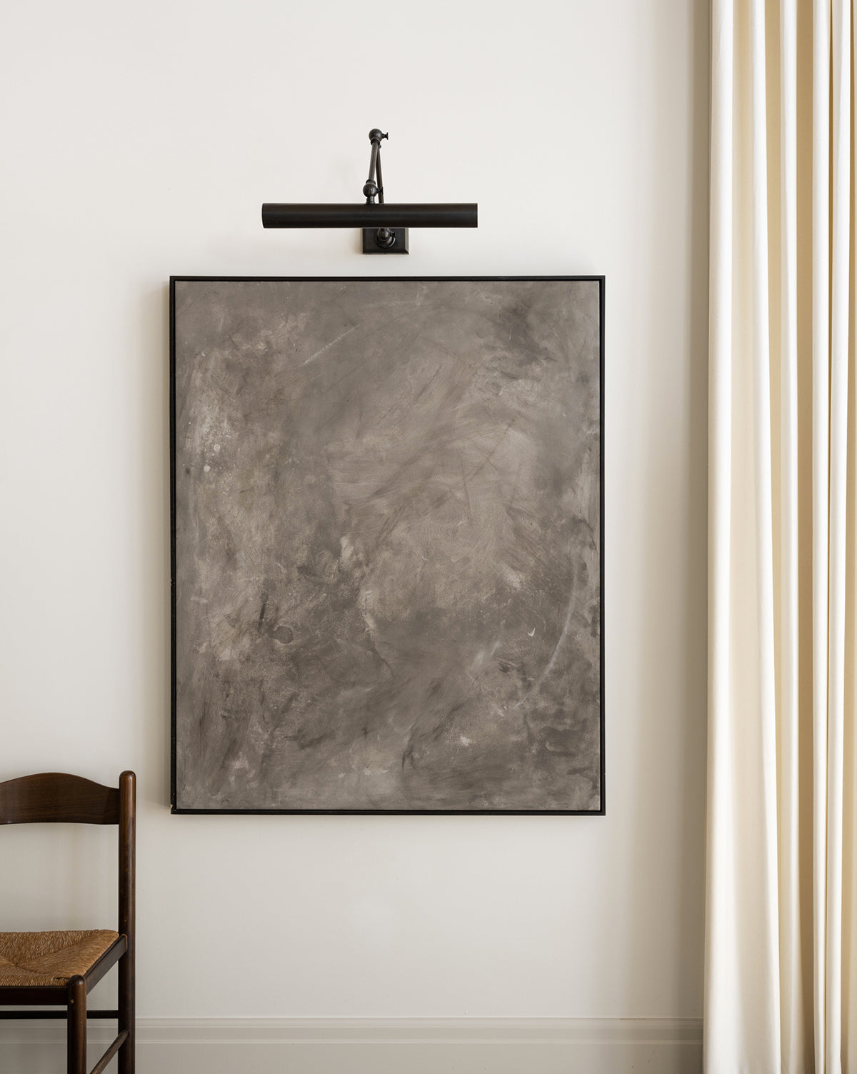 The Nouveau Sepia Abstract large gray giclee print, gallery-wrapped with a black frame, hangs on a white wall under a picture light. A wooden chair with woven seat is on the left and cream-colored curtains are on the right.