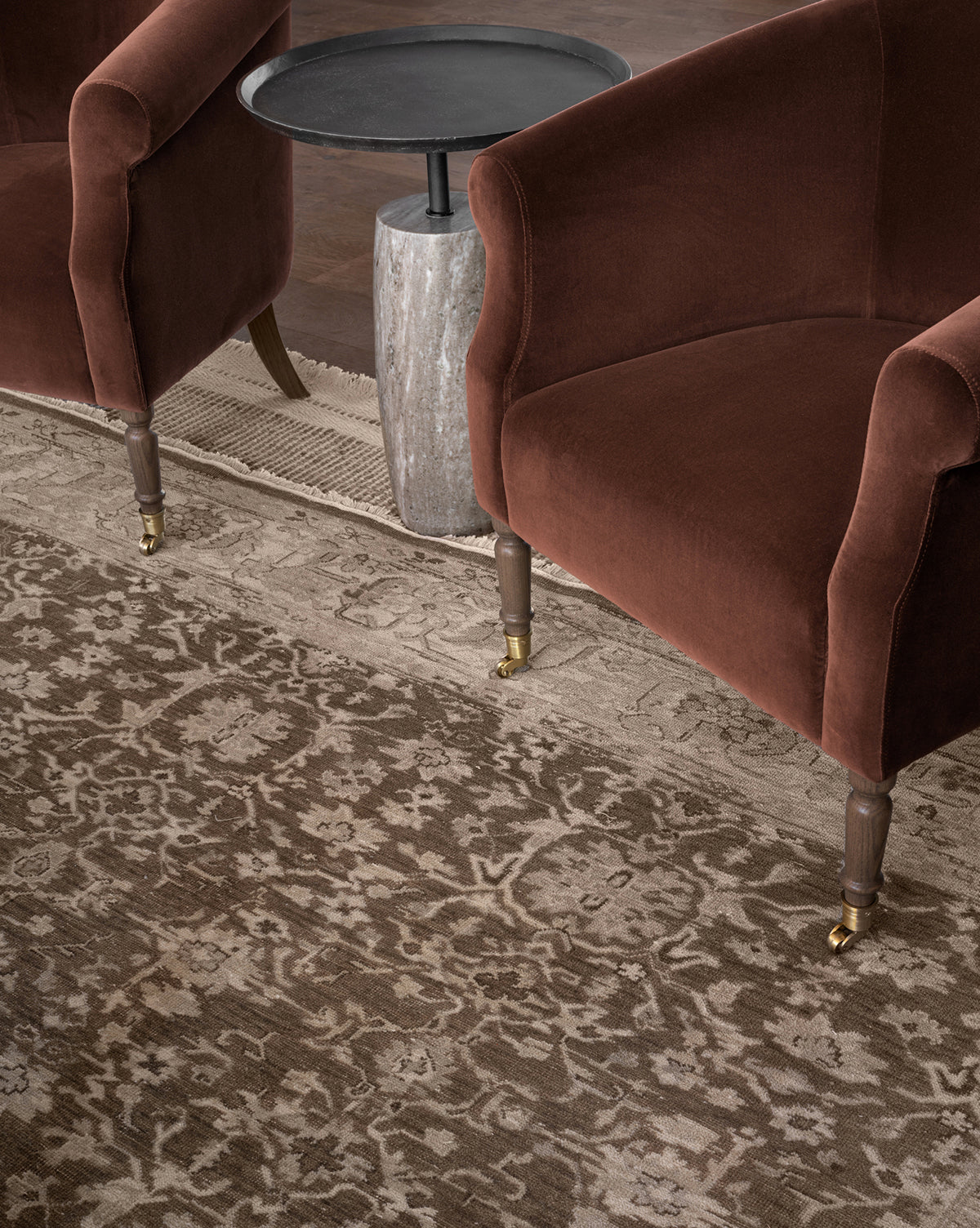 Two Clemence Lounge Chairs (Ready to Ship) with brown designer fabric upholstery, wooden legs, and brass wheels sit on a patterned rug next to a small round black side table with a stone base—ideal for stylish lounge spaces, by McGee & Co.
