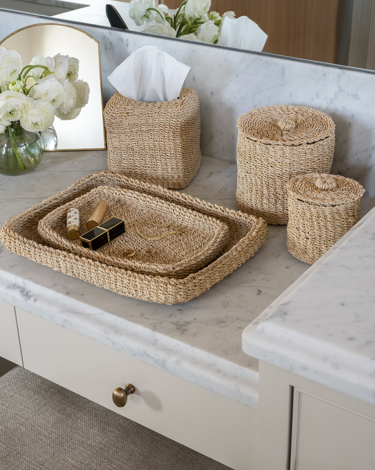 A bathroom countertop with abaca fiber baskets, a Woven Tissue Box Cover, matching tray for lipstick and brush, white flowers in a vase, and a small mirror creates elegant decor on white marble.