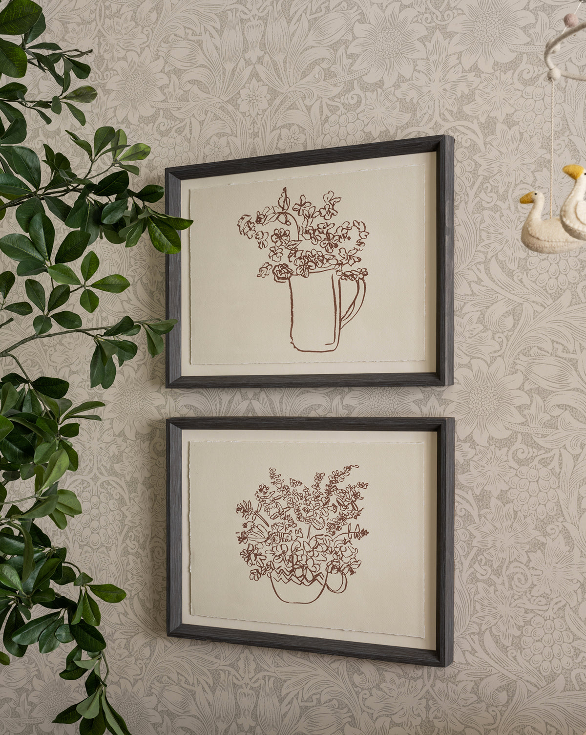 The “Pitcher of Flowers” artwork features vibrant blooms in a vase, framed in charcoal wood grain, hanging on patterned wallpaper beside a leafy plant and an ornament shaped like two swans.
