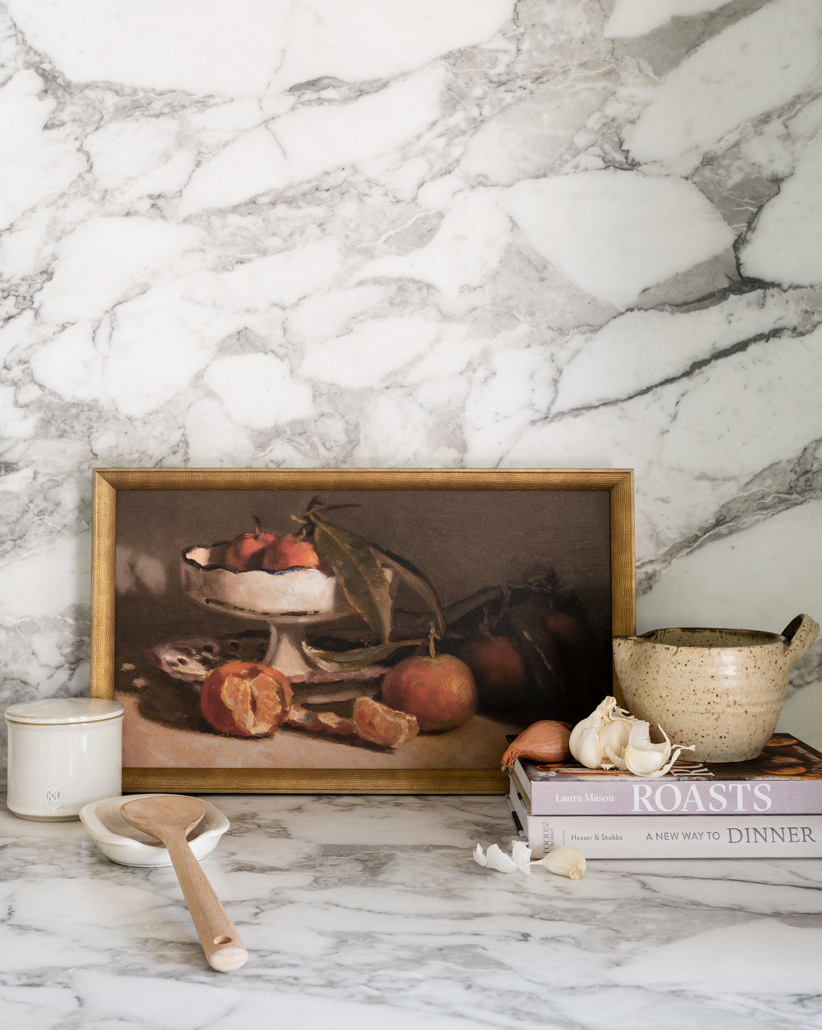 A marble kitchen counter featuring Manderine Still Life by Pascal Giroud, a wooden spoon, a ceramic bowl of orange mandarins, garlic, and stacked cookbooks titled Roasts and A New Way to Dinner.