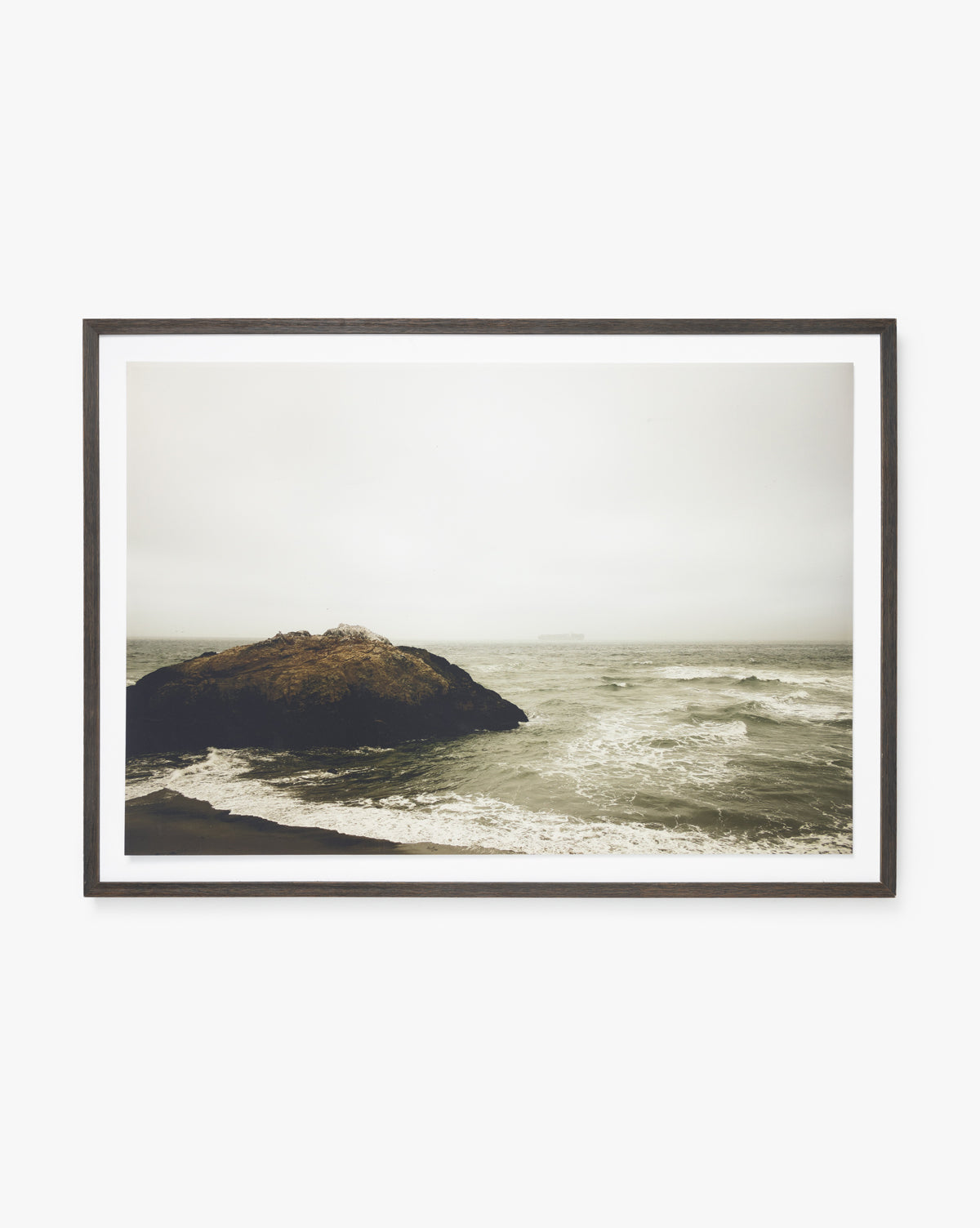 Sunday Fog is a framed giclee print of a rocky outcrop surrounded by waves under an overcast sky, featuring muted tones for a moody coastal vibe—ideal for lovers of foggy coast photography.