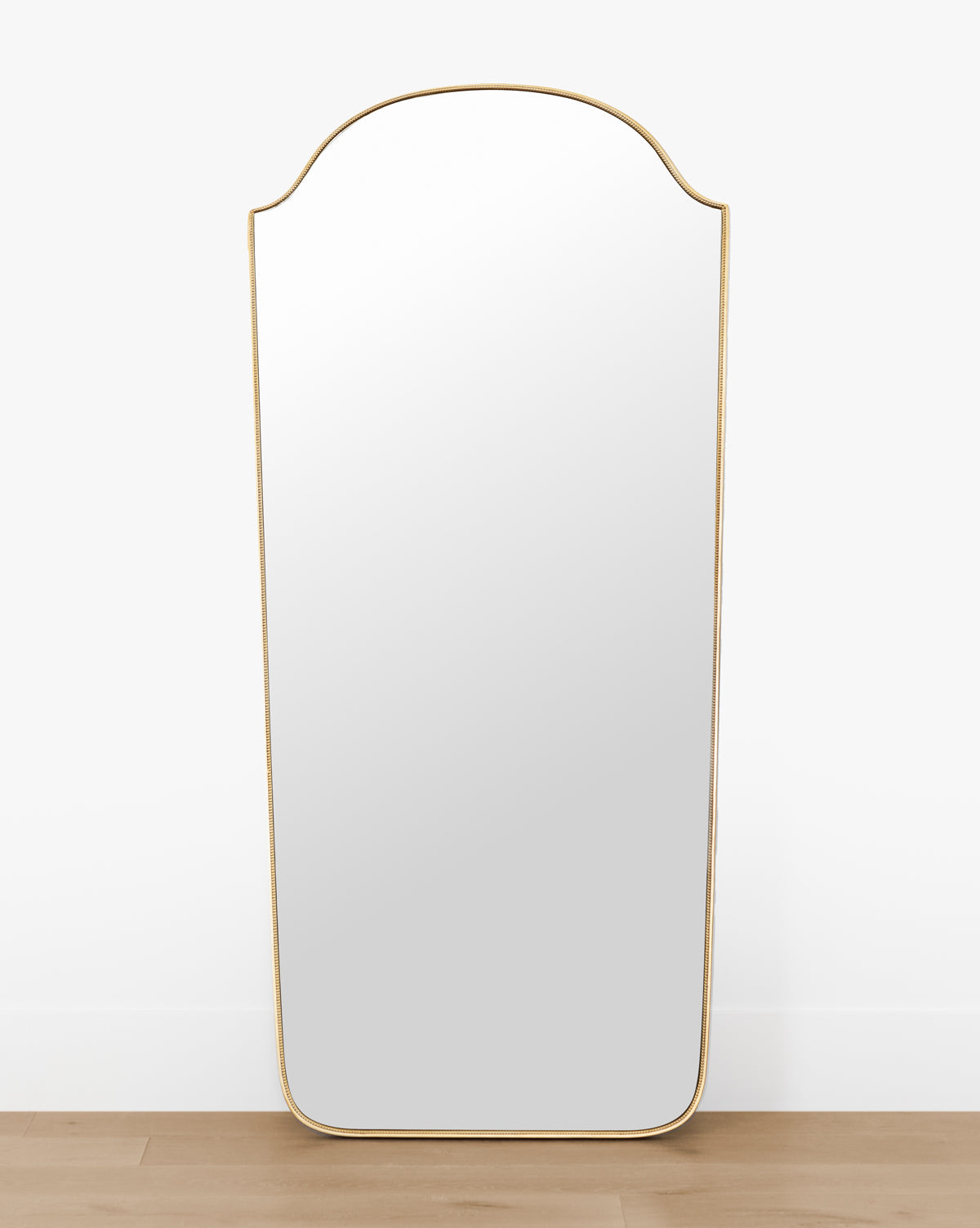 The Sussex Floor Mirror, with its tall arched design and thin antique brass frame, stands against a white wall on a light wood floor. Its rounded top and rectangular bottom corners lend an elegant touch to any space by McGee & Co.