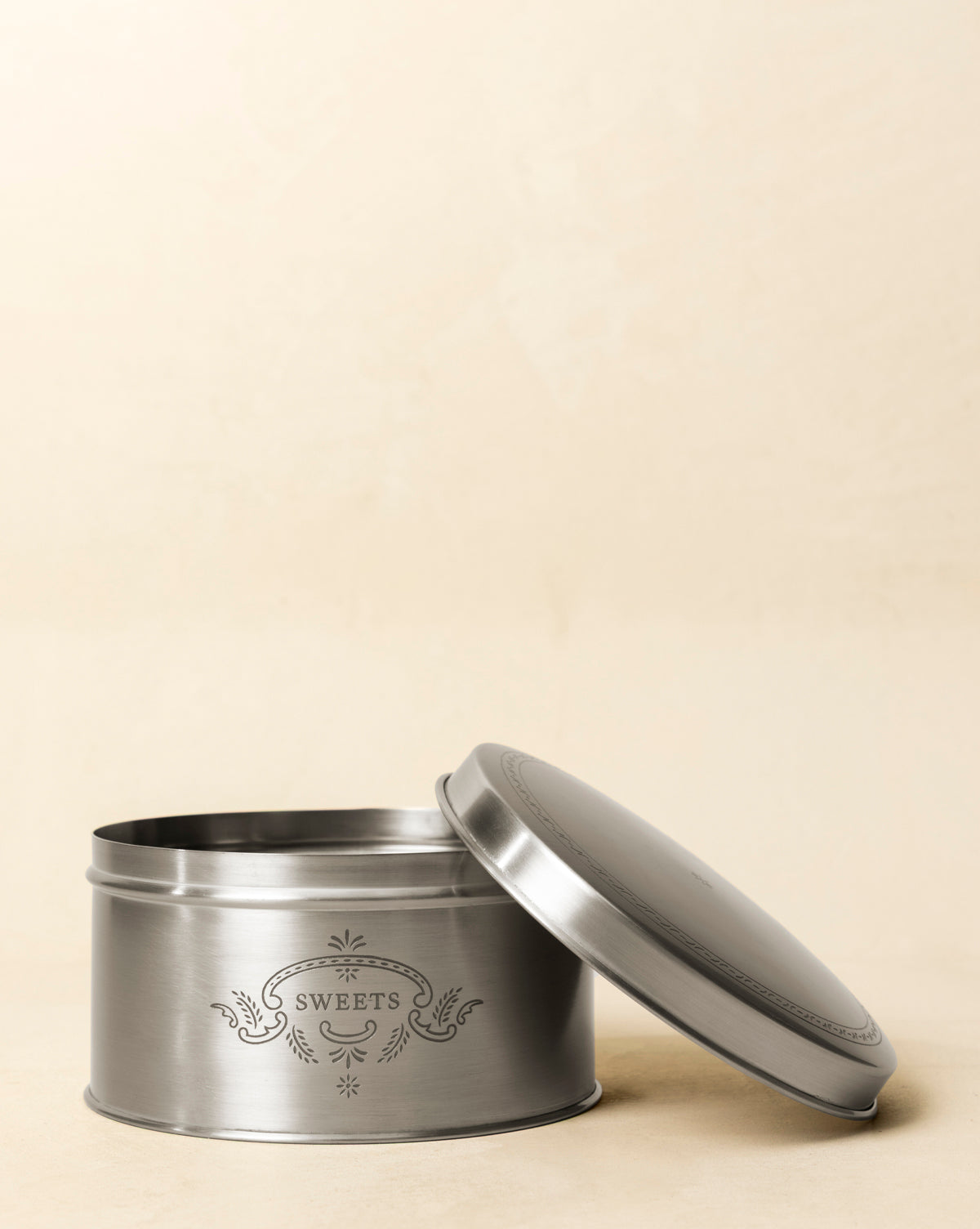 The Sweets Cookie Tin, a round silver metal container with a decorative label, sits open with its lid angled beside it, adding vintage charm against a plain beige background, by McGee & Co.