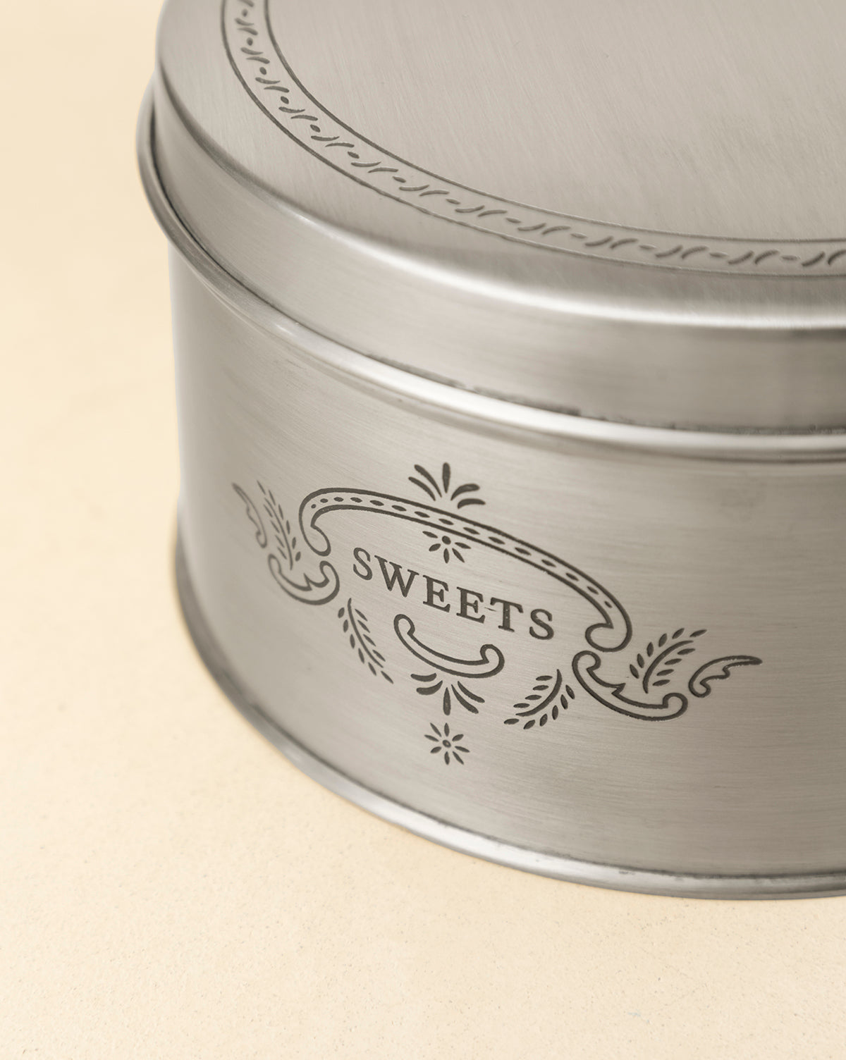 The Sweets Cookie Tin is a round, silver container with vintage-inspired decorative patterns and "SWEETS" engraved on the side, ideal for gifting holiday treats. It is displayed on a light beige surface, by McGee & Co.