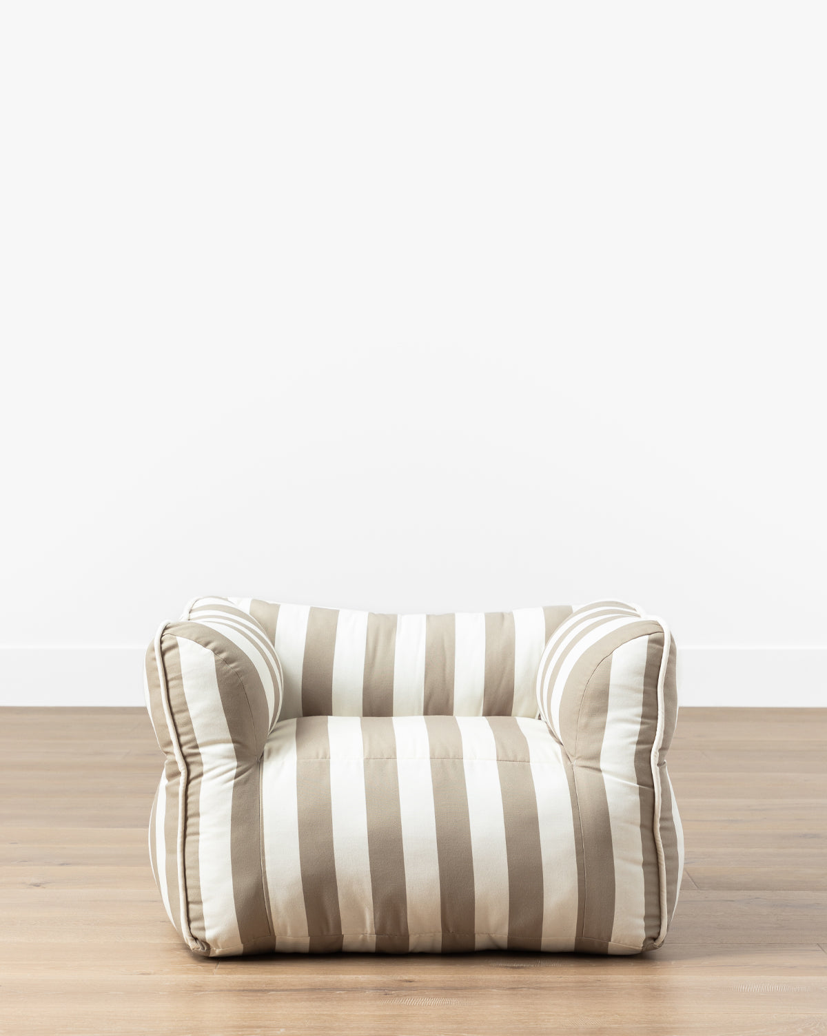 The Tan Striped Kids Chair, featuring beige and white horizontal stripes, sits on a wooden floor against a plain white wall—ideal for bringing charm to a child’s room, by McGee & Co.
