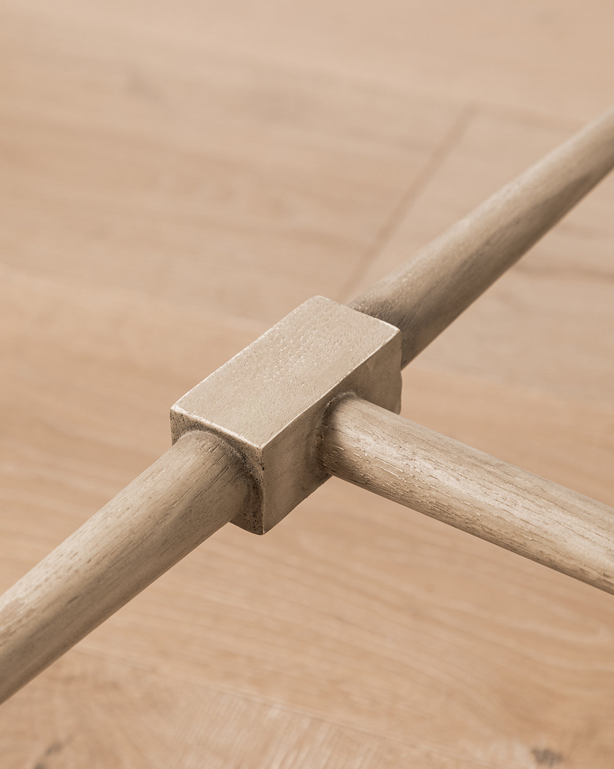 Close-up of intersecting solid white oak rods joined by a rectangular metallic connector, showcasing the craftsmanship and modern design of the Tavin Side Table, with a softly blurred wood surface in the background.