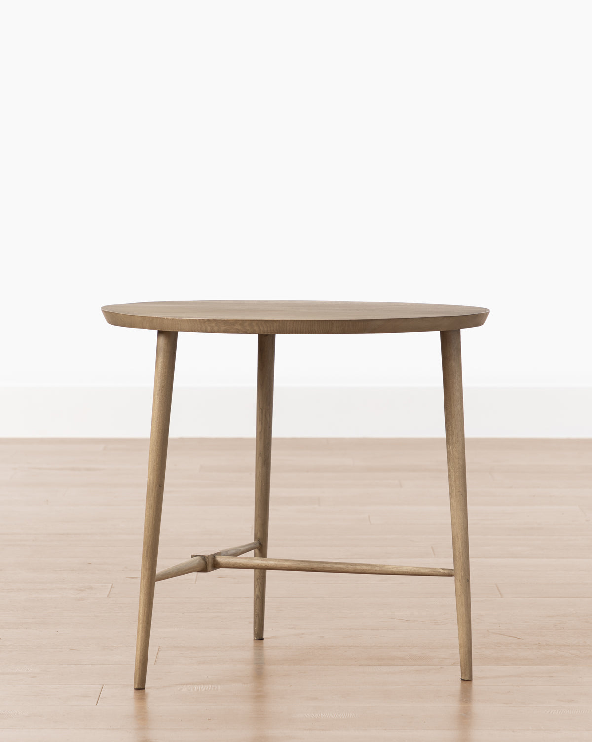 The Tavin Side Table, featuring a round solid white oak top, three slim legs, and a supportive crossbar, is placed on a light wood floor against a plain white background.