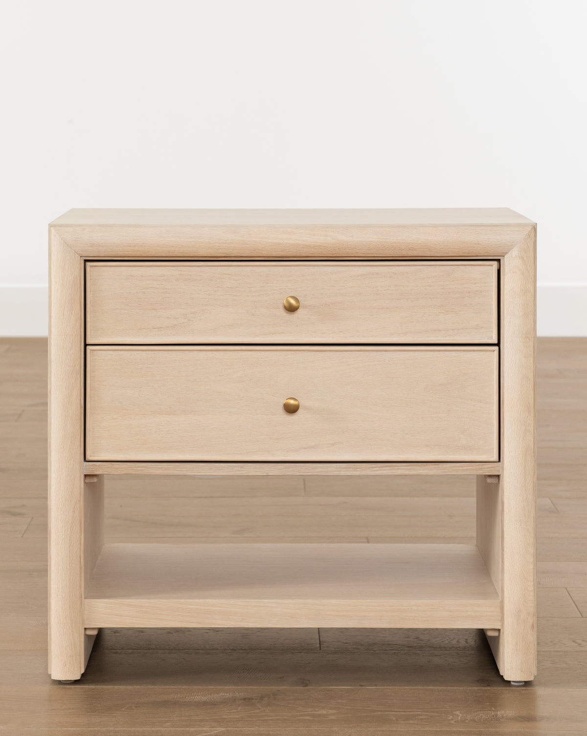 The Tellerbeck Nightstand showcases a light, bleached oak finish with two drawers accented by gold knobs and an open lower shelf, ideal for bedroom styling, by McGee & Co. Displayed on a wooden floor with a plain white backdrop.