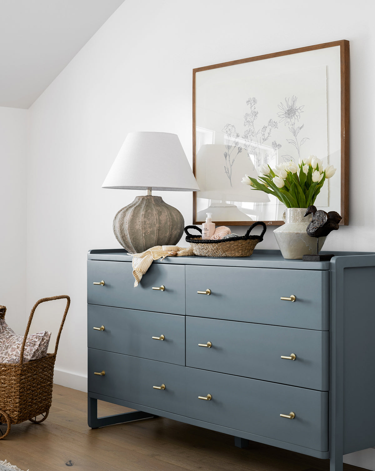 A blue dresser with gold knobs holds a ceramic lamp, a Petites Fleurs framed print, white tulips in a vase, and a wicker basket with eggs. Another woven basket is placed on the floor next to the dresser.