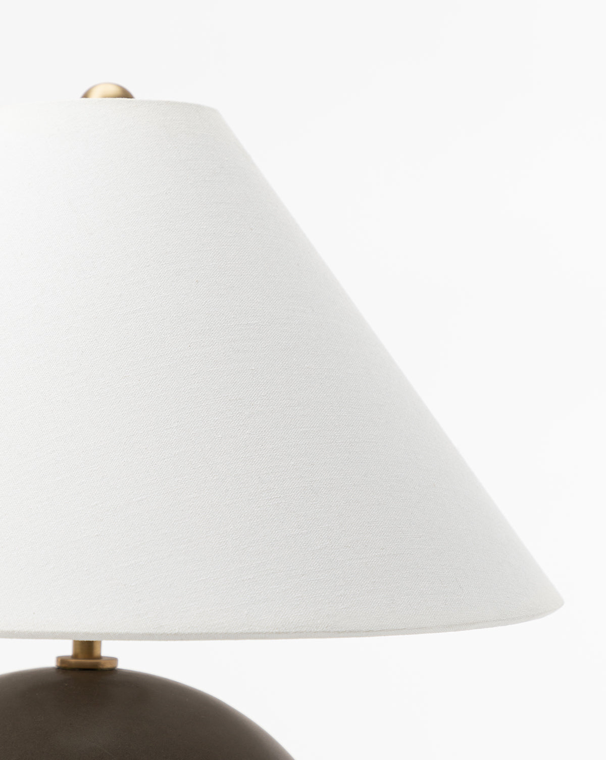 Close-up of the Tess Ceramic Table Lamp featuring a white, cone-shaped lampshade with a brass finial on top, set against a plain white background with the lower part of its ceramic base partially visible, by McGee & Co.