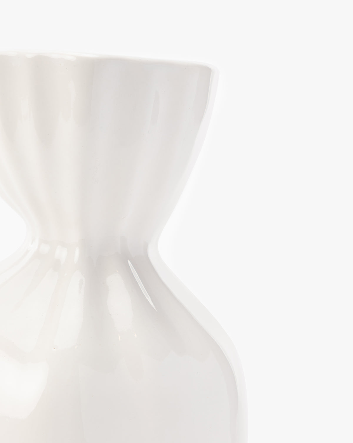Close-up of the Thessaly Vase, a glossy white modern piece with a unique hourglass shape, partially visible against a plain white background and featuring soft reflections on its smooth surface.