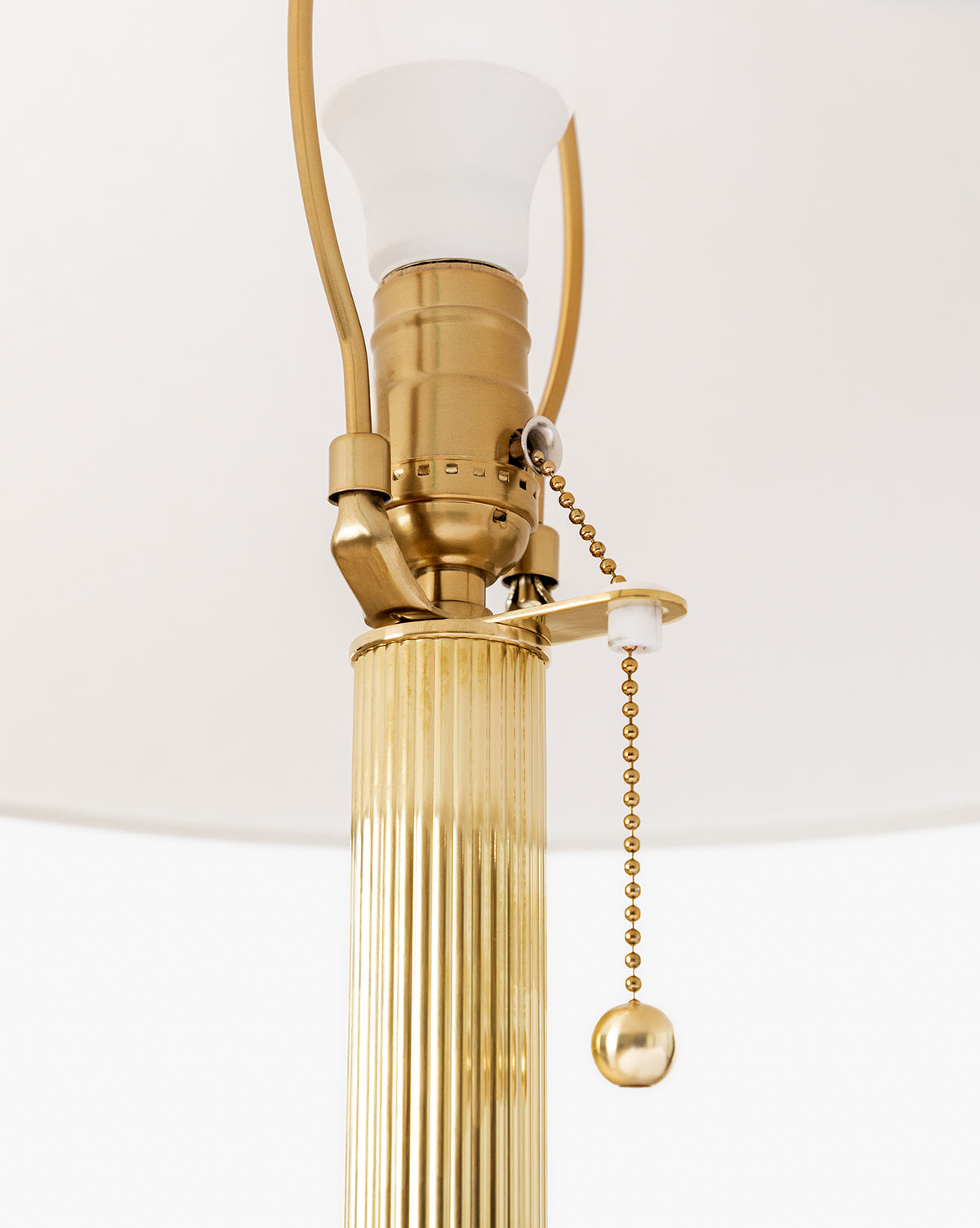 Close-up of the Townes Floor Lamp, featuring a reeded brass base, pull chain with round end, frosted white bulb socket, and partially covered by a white linen shade by McGee & Co.