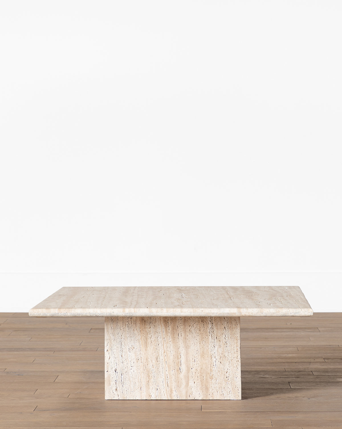 The Travertine Coffee Table features a rectangular, handmade slab top and a wide pedestal base, shown here on a wooden floor against a white wall, by McGee & Co.