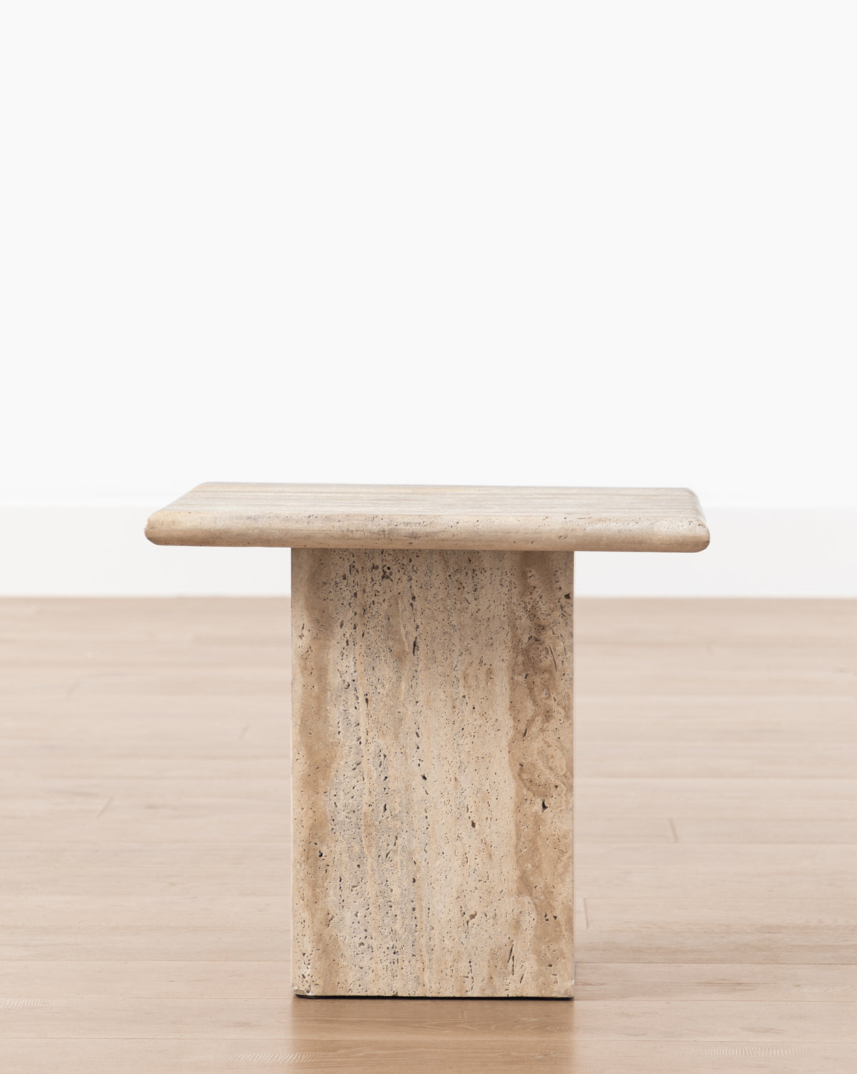 The Travertine Side Table features a minimalist rectangular design with a thick travertine marble top and a single wide pedestal base, shown on a light wood floor against a plain white wall, by McGee & Co.