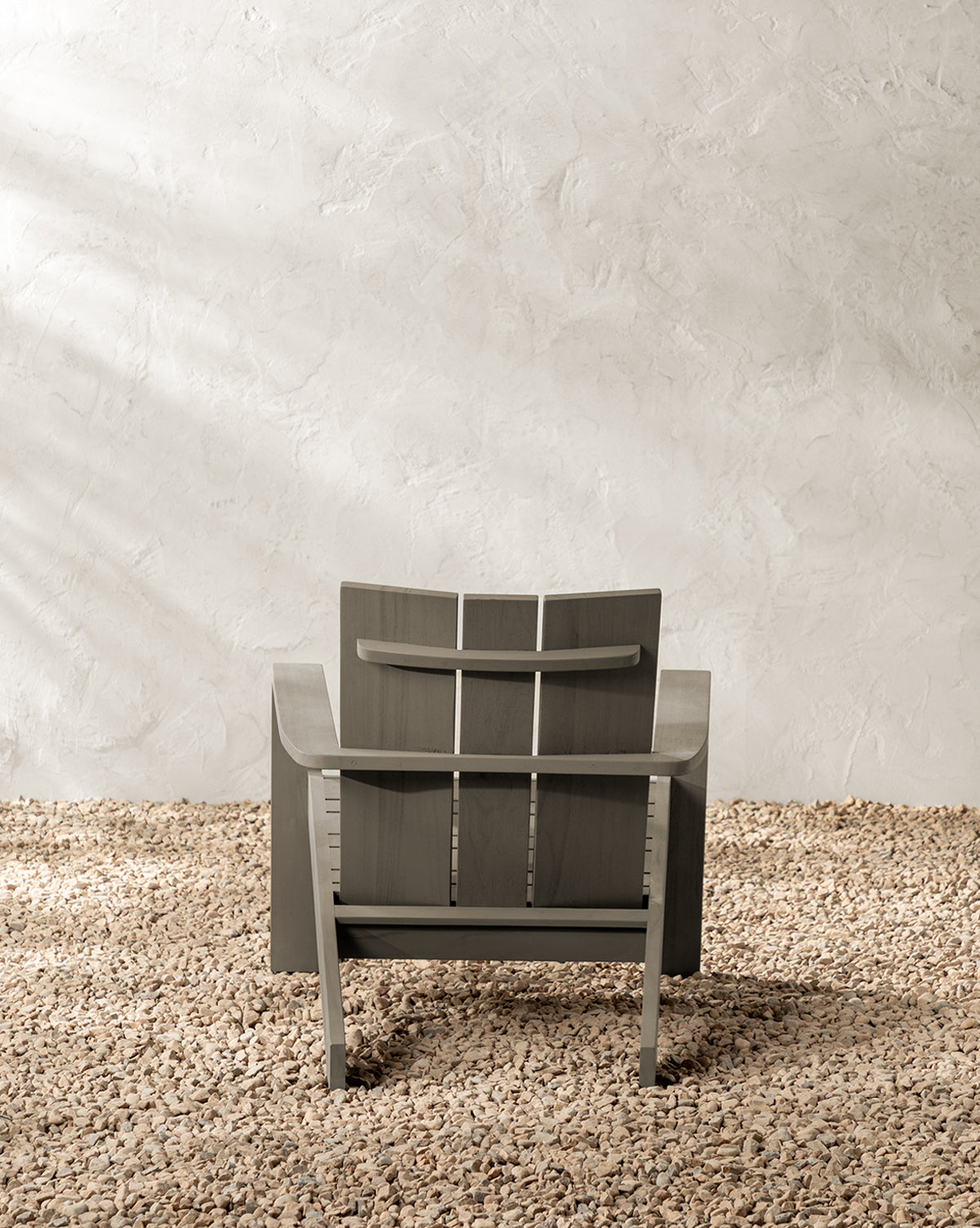 The Triby Gray Teak Outdoor Lounge Chair, featuring a slatted backrest, rests on small light brown pebbles against a textured off-white wall, with soft natural light creating gentle shadows by McGee & Co.