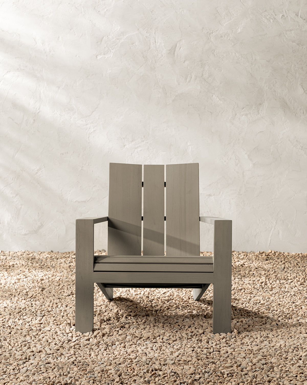 The Triby Gray Teak Outdoor Lounge Chair features wide armrests and a slatted backrest, placed on light pebbles against a textured beige wall—ideal for enhancing any patio space by McGee & Co.