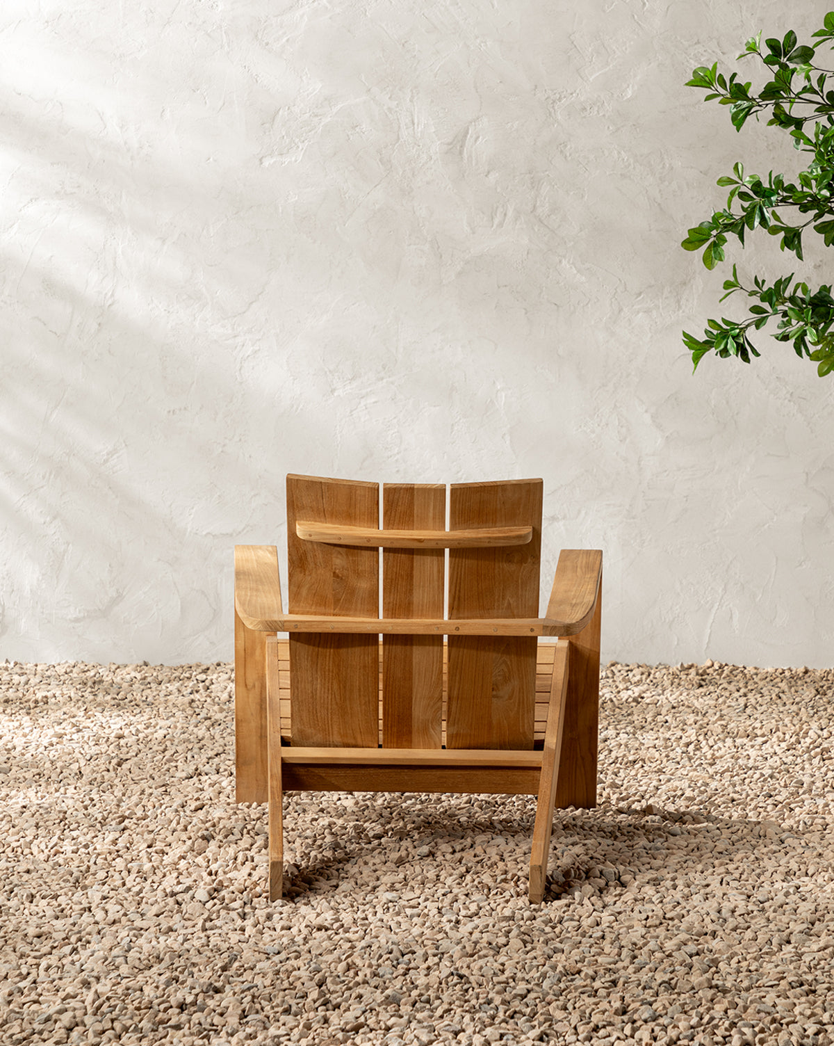 The Triby Teak Outdoor Lounge Chair with wide armrests rests on pebbles before a textured white wall, while a green leafy plant on the right adds to the cozy patio vibe, by McGee & Co.