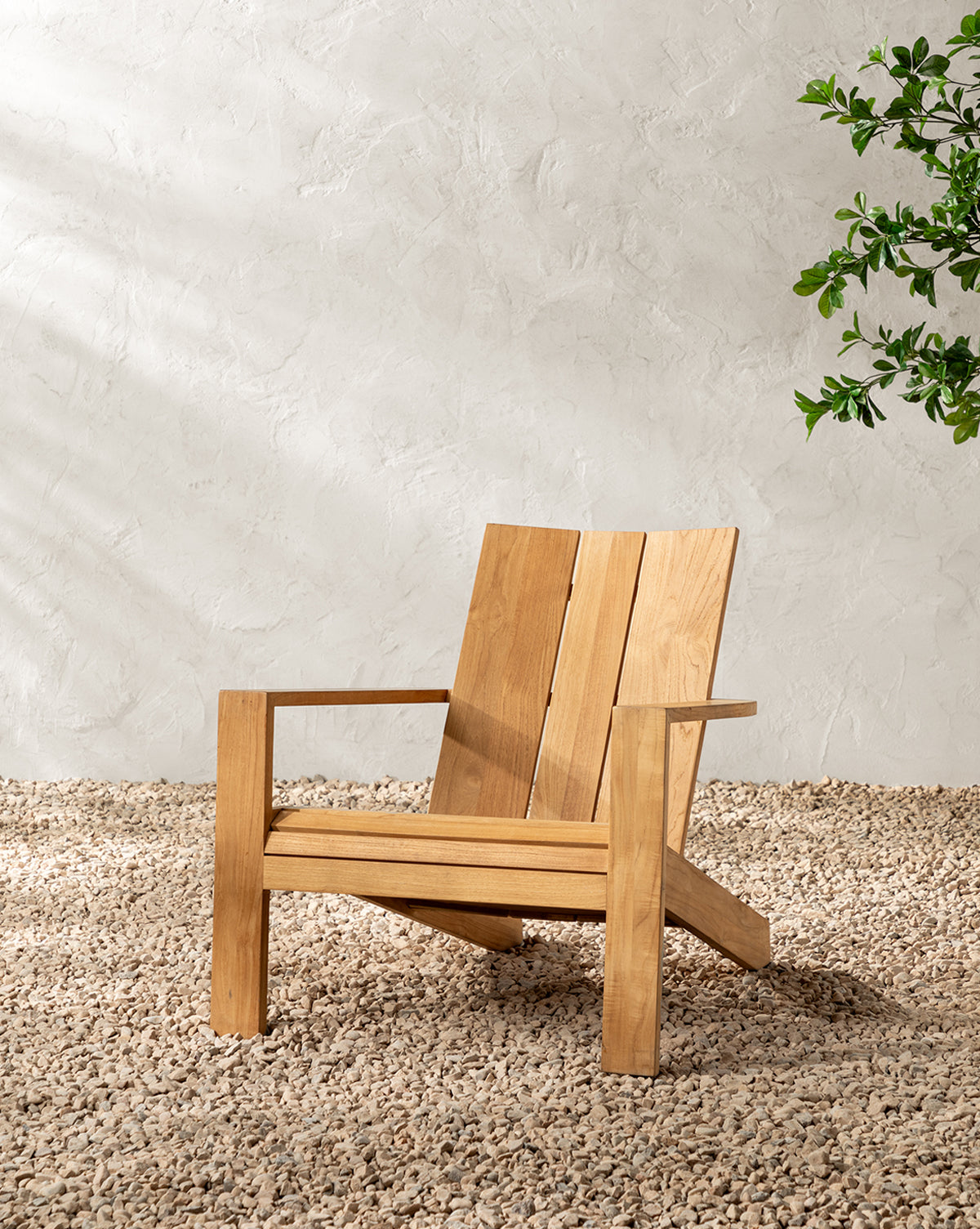 The Triby Teak Outdoor Lounge Chair rests on small pebbles by a light textured wall, crafting a stylish patio retreat, while green leaves from a nearby plant appear in the top right corner by McGee & Co.