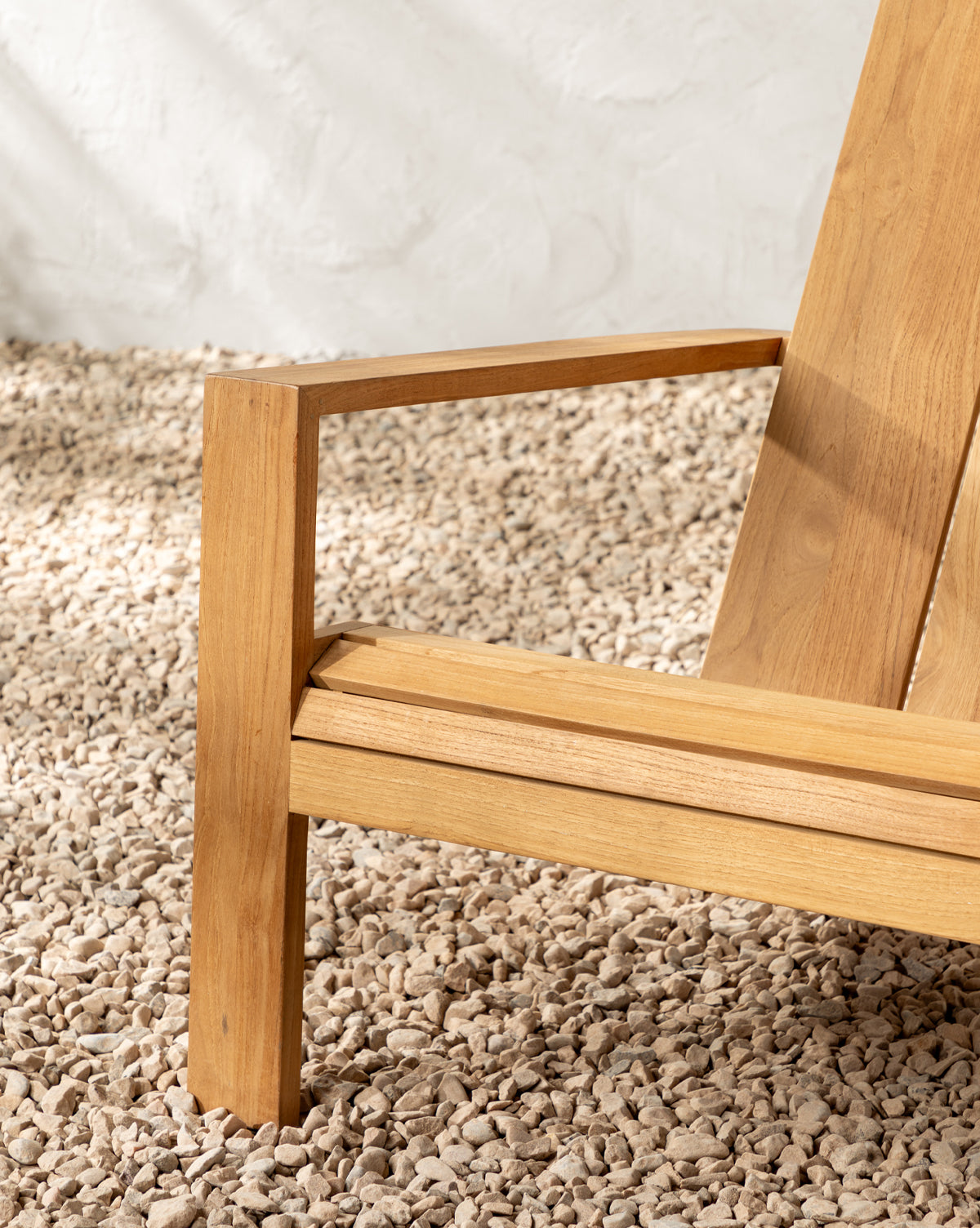 A close-up of the Triby Teak Outdoor Lounge Chair, featuring clean, modern lines and set on light pebbles against a textured white wall—perfect for any all-weather patio setup, by McGee & Co.