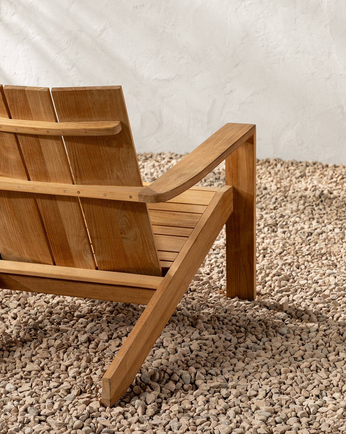 A close-up of the Triby Teak Outdoor Lounge Chair’s back and armrest on light gravel, set against a textured white wall—this all-weather chair is perfect for patios and elevates any outdoor space, by McGee & Co.