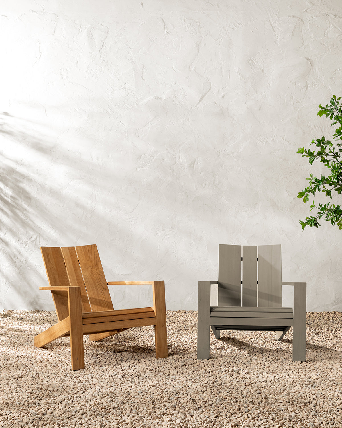 Two modern patio chairs, including the Triby Teak Outdoor Lounge Chair and a gray all-weather chair, sit on gravel against a textured white wall. A leafy plant to the right and sunlight create gentle shadows, enhancing the inviting scene by McGee & Co.