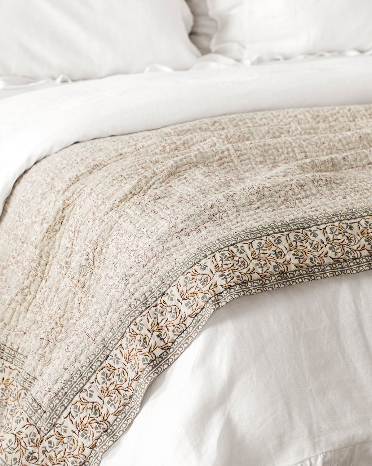 A close-up of a bed with white linens, draped with the Tulare Block Print Quilt—beige with a floral border and block print design, by McGee & Co.