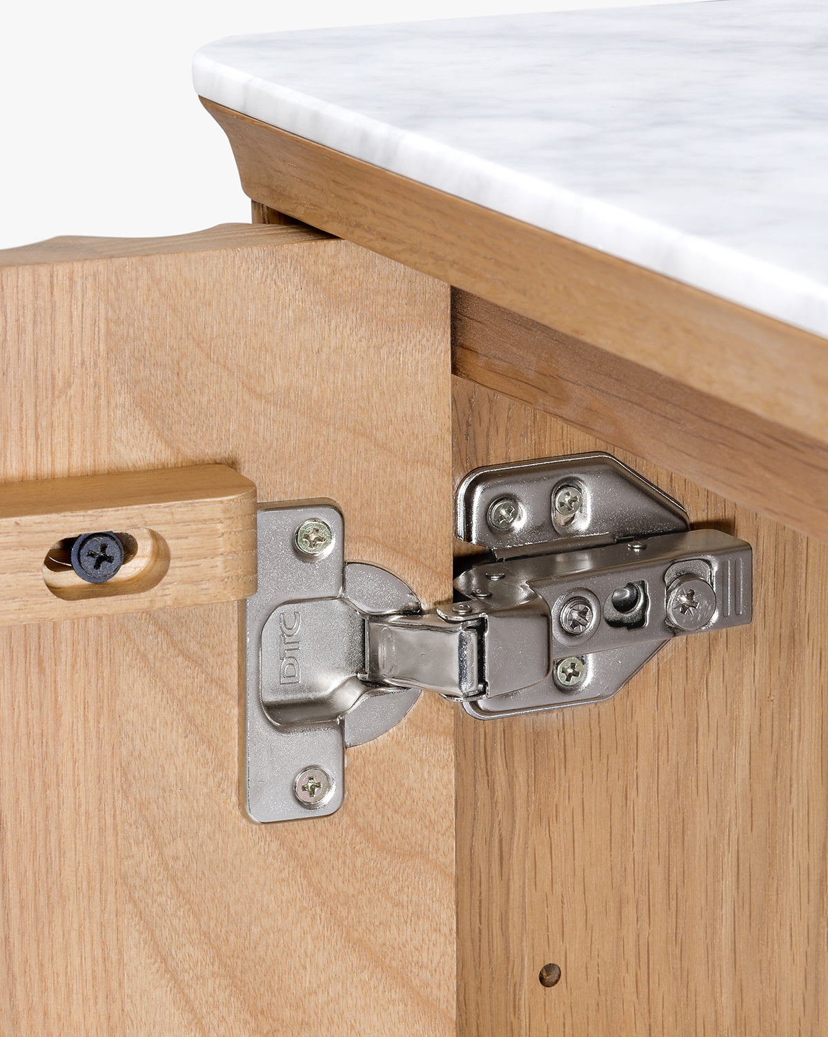 A close-up of a silver metal hinge on the inside of a wooden Vale Sideboard door, with its marble top console and white countertop visible above, by McGee & Co.