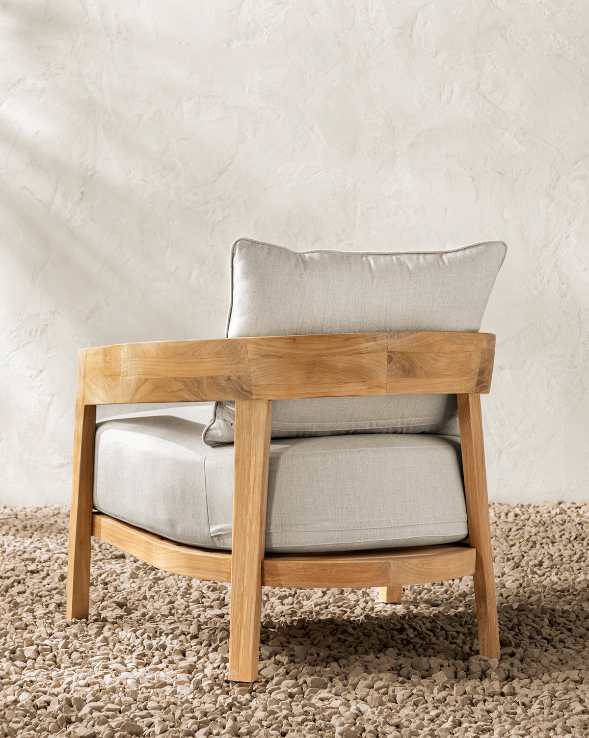 The Vento Teak Outdoor Lounge Chair with light beige cushions and rounded wooden arms sits on a textured stone floor against a pale wall, offering a minimalist, modern look ideal for stylish outdoor lounge arrangements by McGee & Co.