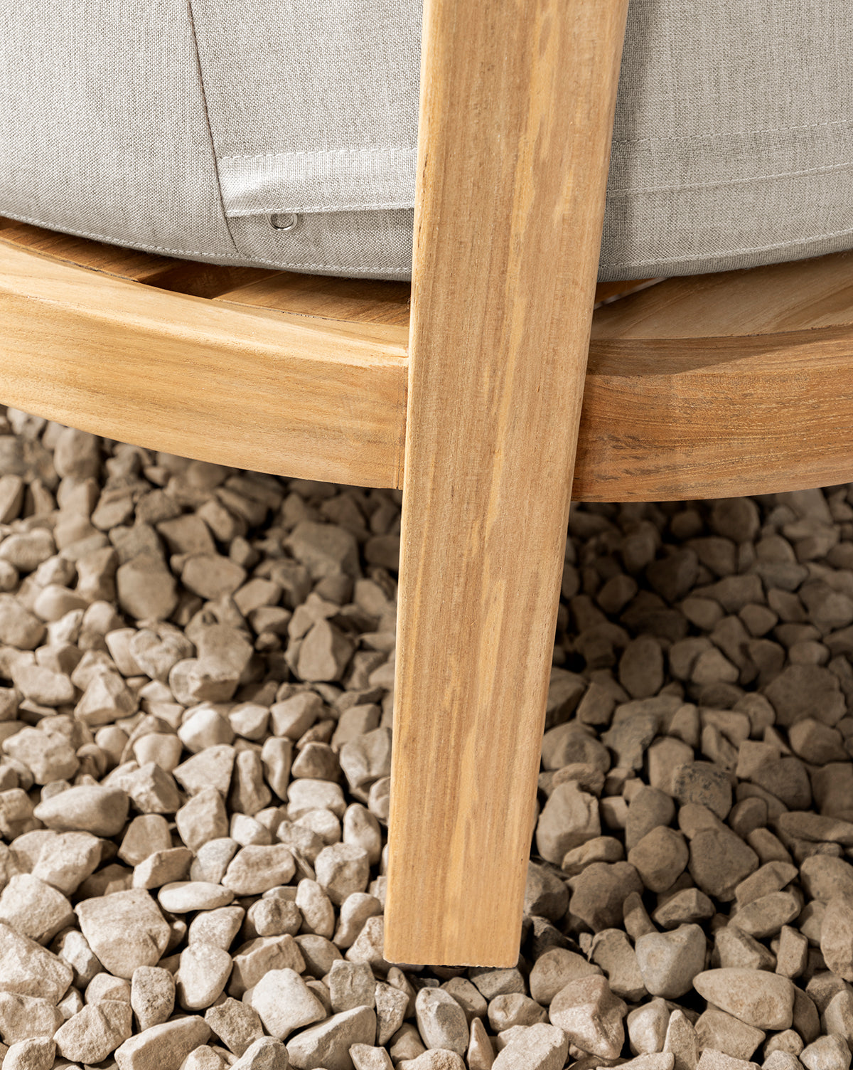 Close-up of a Vento Teak Outdoor Lounge Chair leg on small, light-colored gravel, with part of a light gray cushion visible above the wooden frame, by McGee & Co.
