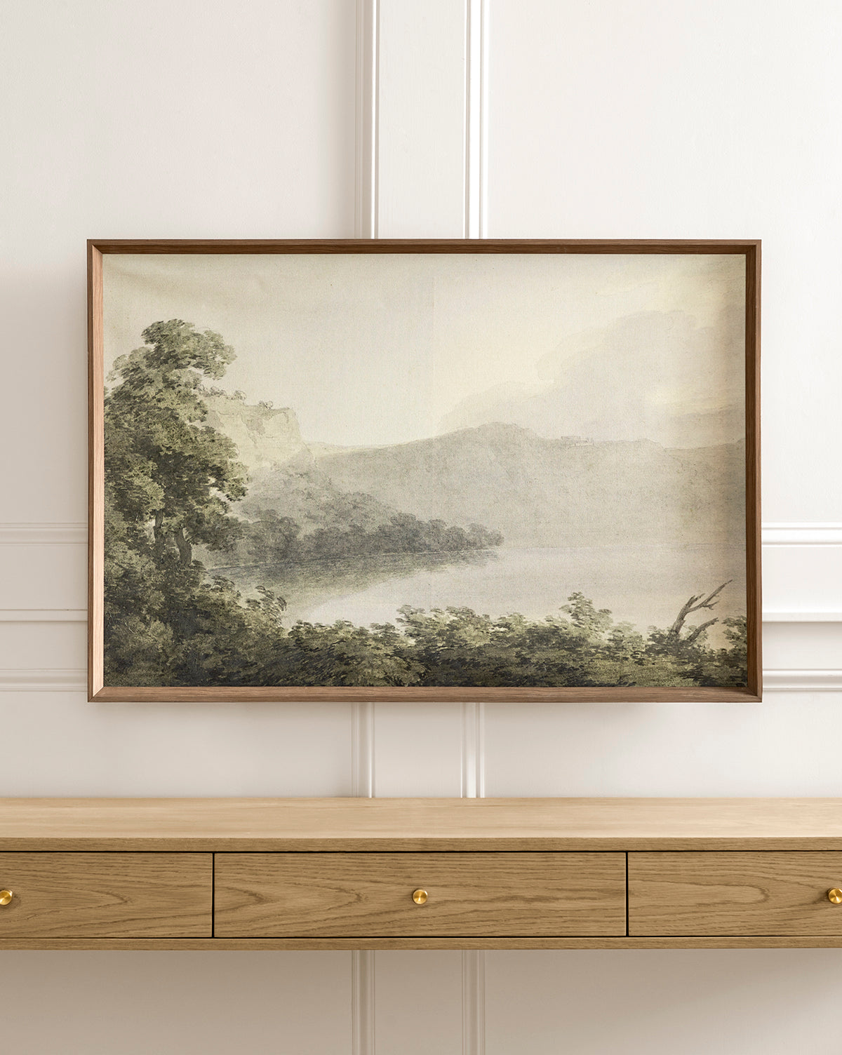 The Verde Landscape giclee print, with misty hills and a tranquil lake among green trees, hangs above a light wood three-drawer console table with gold knobs.