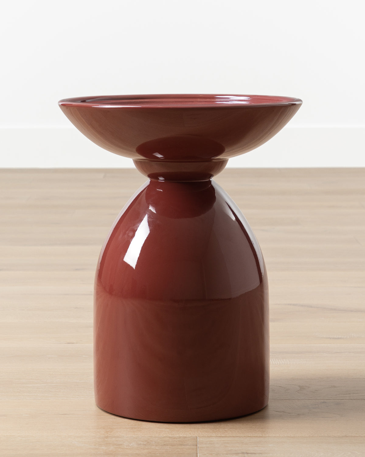 The Visby Side Table features a modern, glossy cherry mahogany finish, a wide shallow top, and a rounded base, displayed on a light wooden floor against a plain white wall by McGee & Co.