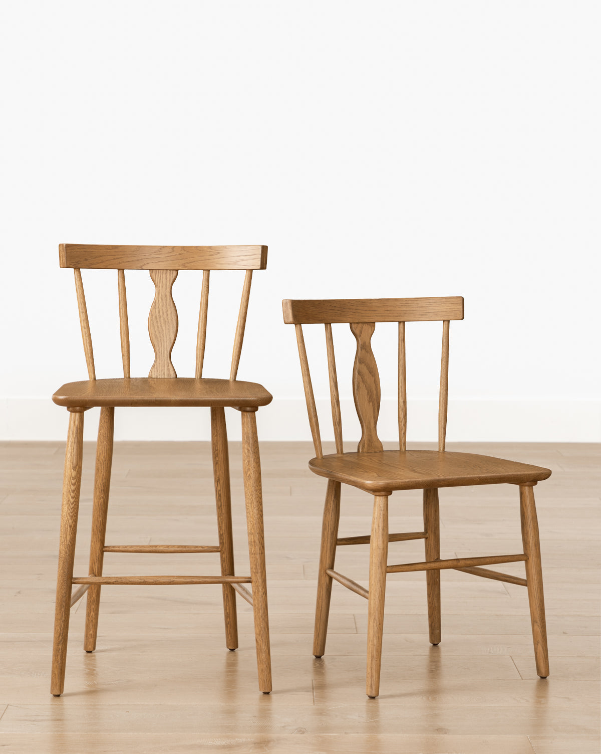 Two Whittaker Chairs—one bar stool and one standard—featuring solid oak construction, spindle backs, and curved seats, are displayed on a light wood floor against a white wall, ideal for elevating any dining space by McGee & Co.